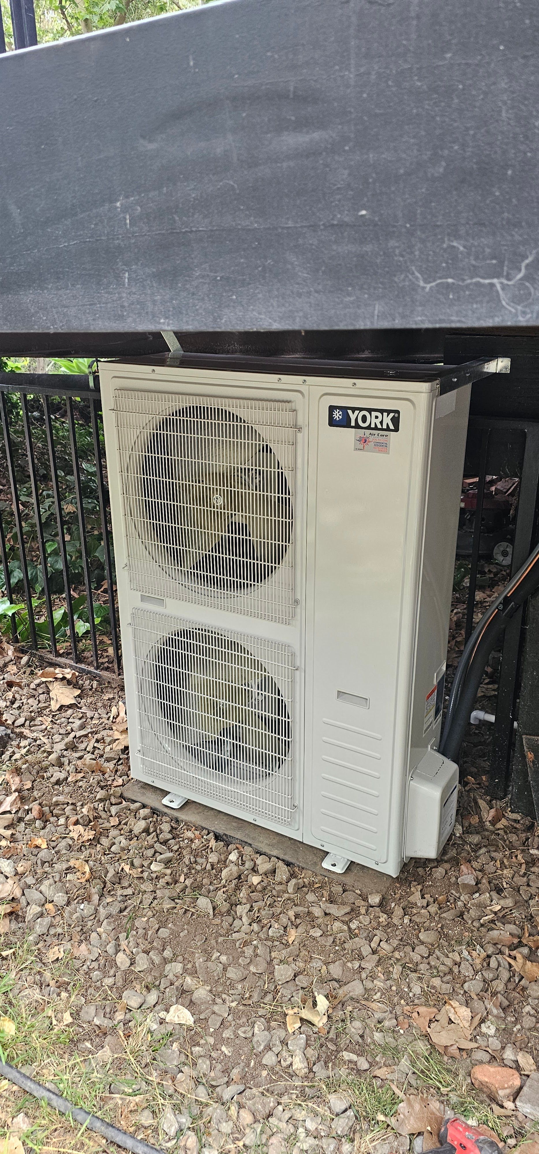Outdoor air conditioner unit beside a dark wall, with two fans and white casing among gravel and foliage