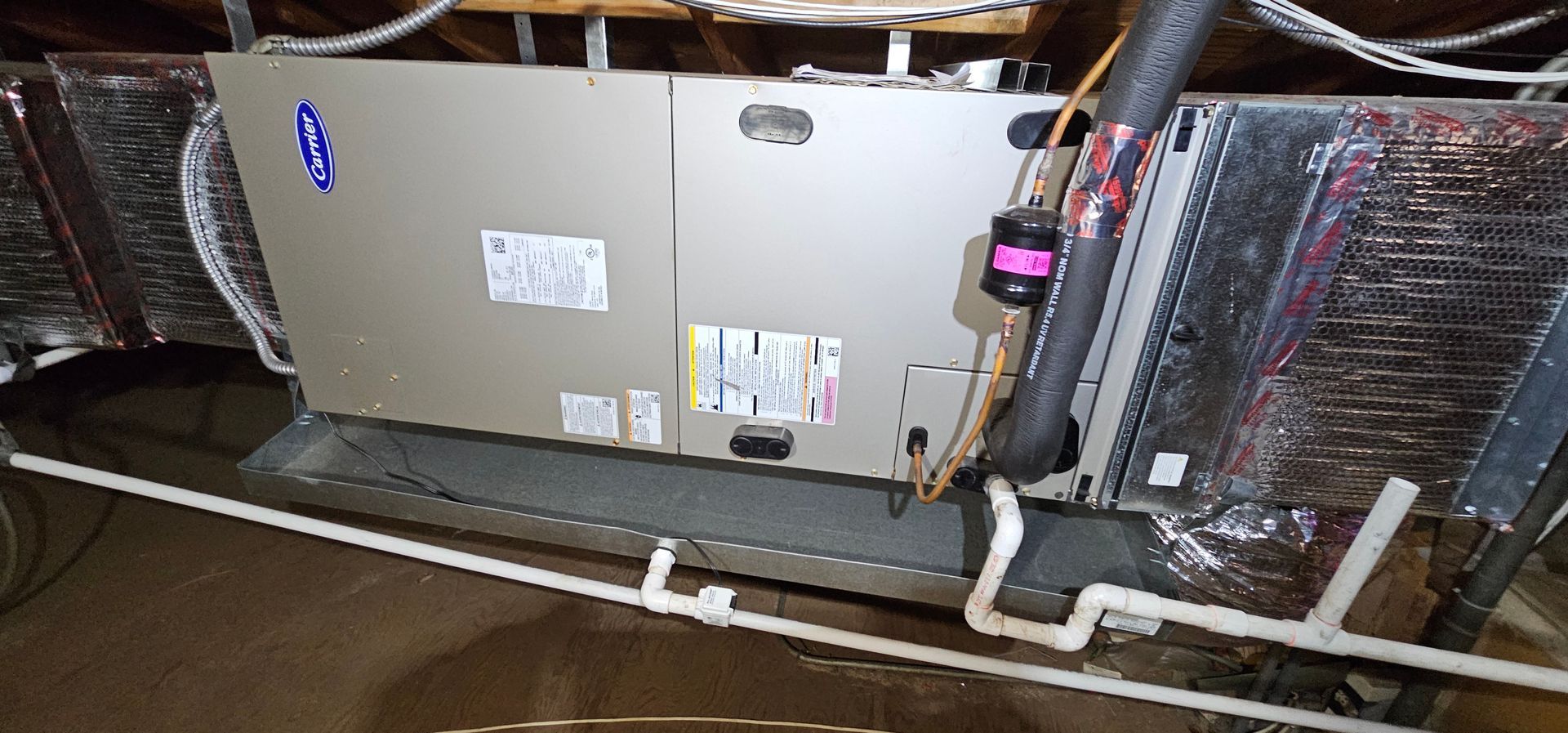 Large HVAC furnace unit in an attic, with insulated ducts and exposed pipes around it.