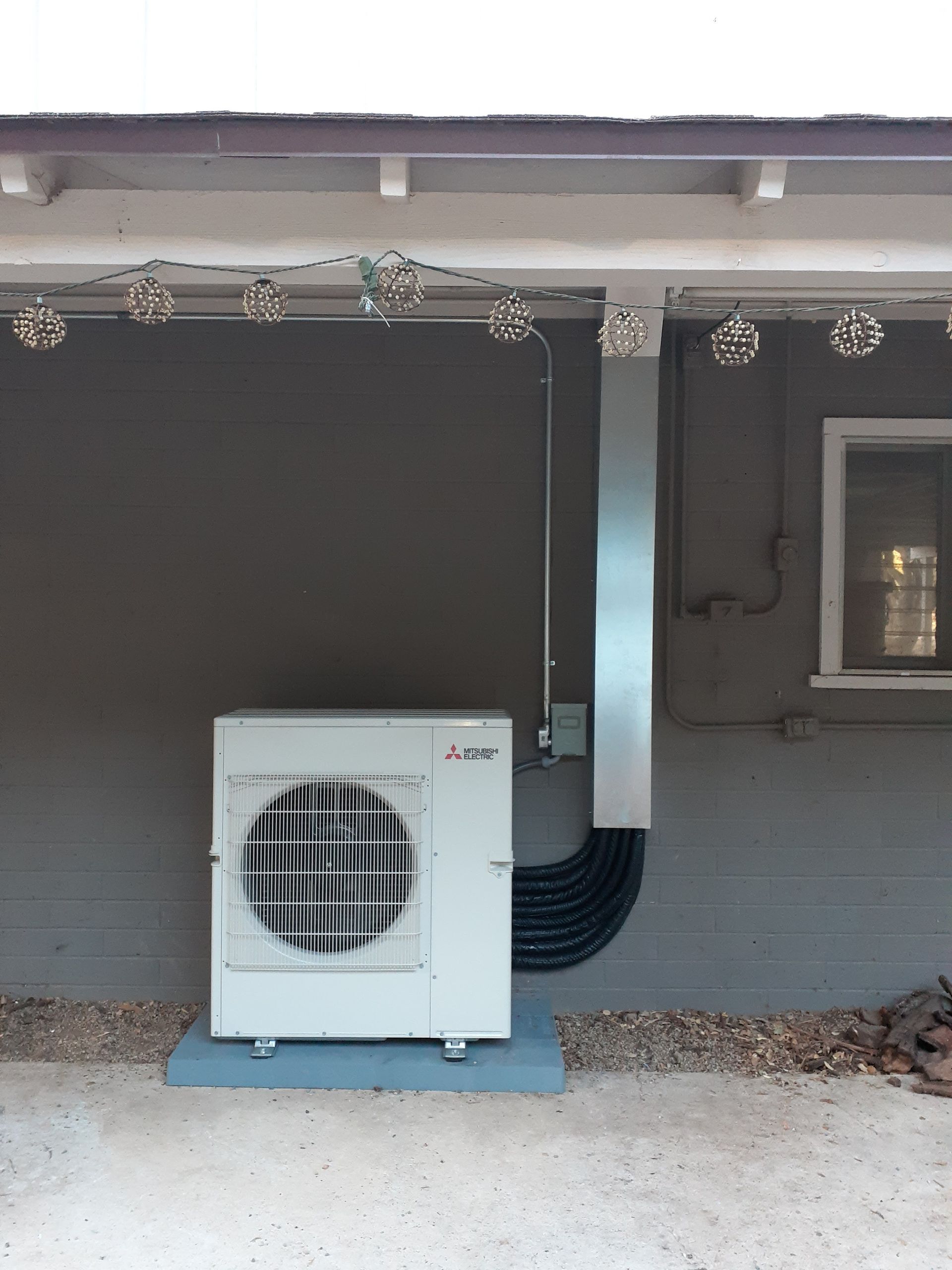 Outdoor heat pump unit beside a gray house wall, with black insulated tubing and a small window nearby