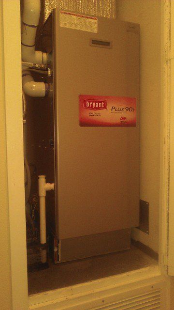 Tall beige boiler/water heater in a narrow utility closet, with a red label on the front.