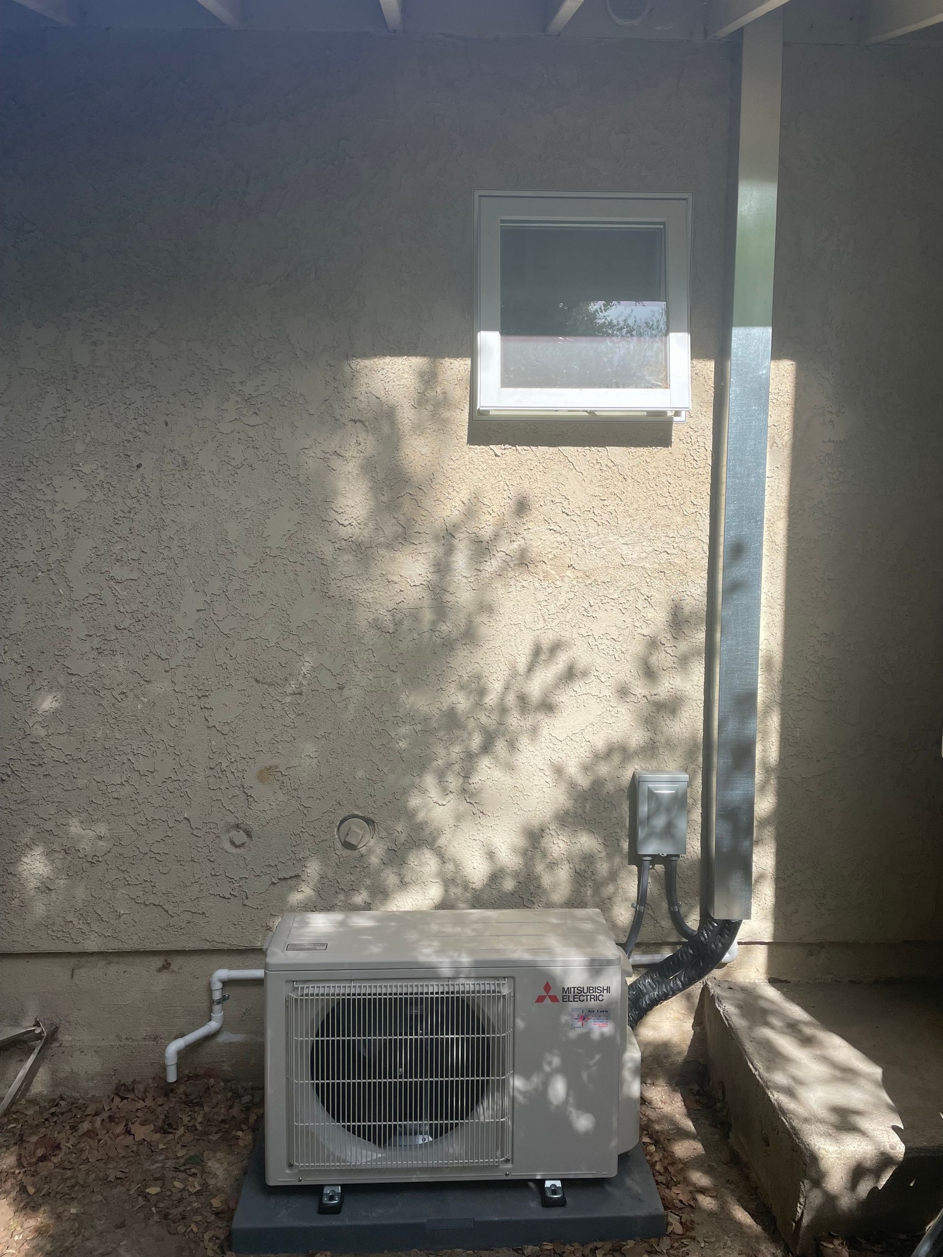 Outdoor air-conditioning unit beside a stucco wall under a small window, with sunlight and shadows.