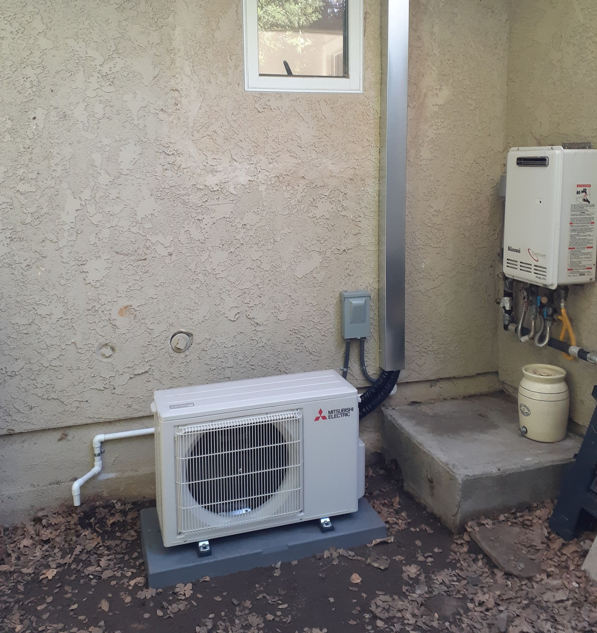 Outdoor HVAC condenser beside a stucco wall, with utility boxes and pipes on a concrete pad.