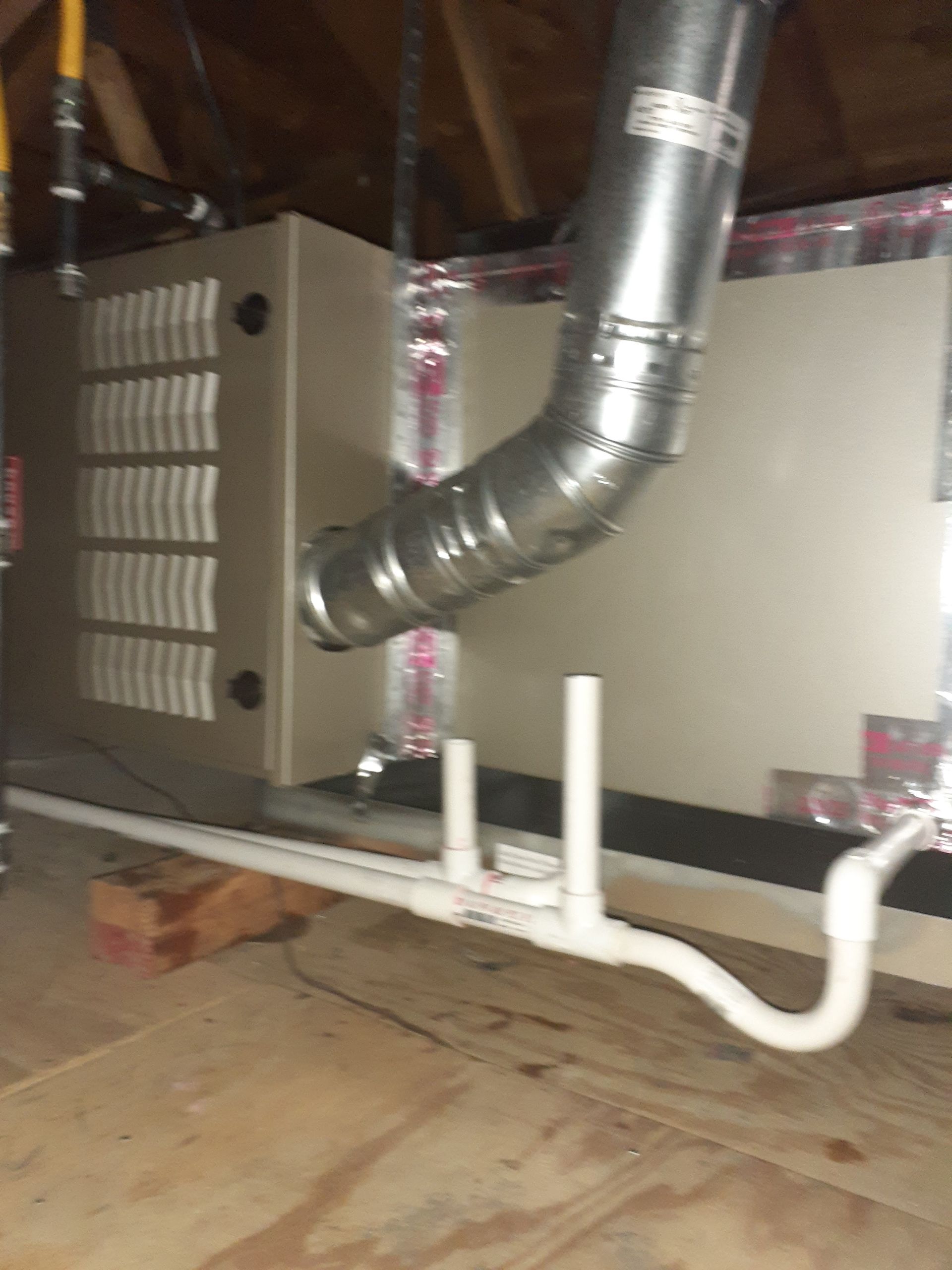 Attic HVAC unit with silver ducting and white PVC pipes on a plywood floor.