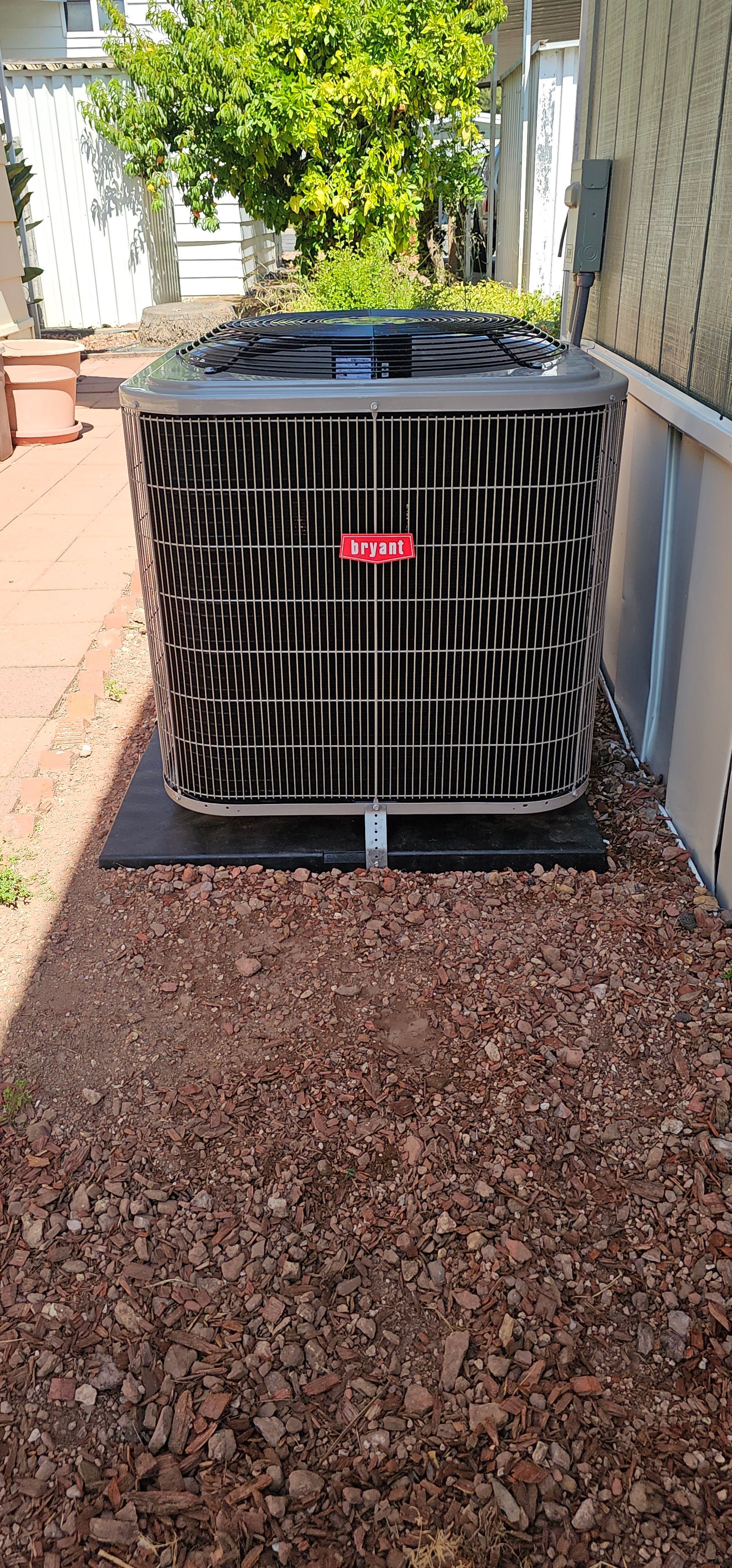 Large black HVAC unit outdoors on a gravel path beside a house and plants.