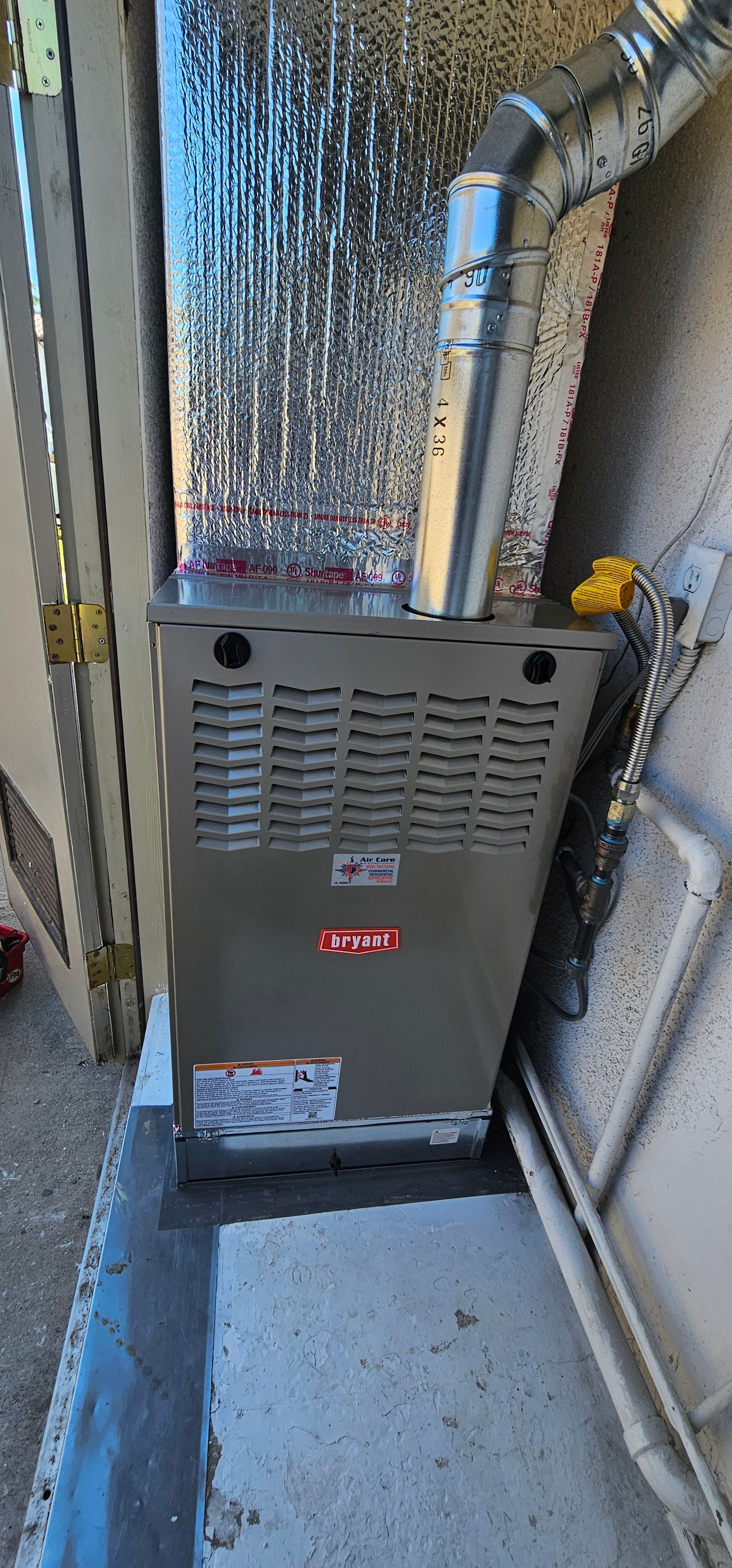 Residential furnace in a narrow utility alcove with silver ductwork and concrete floor.