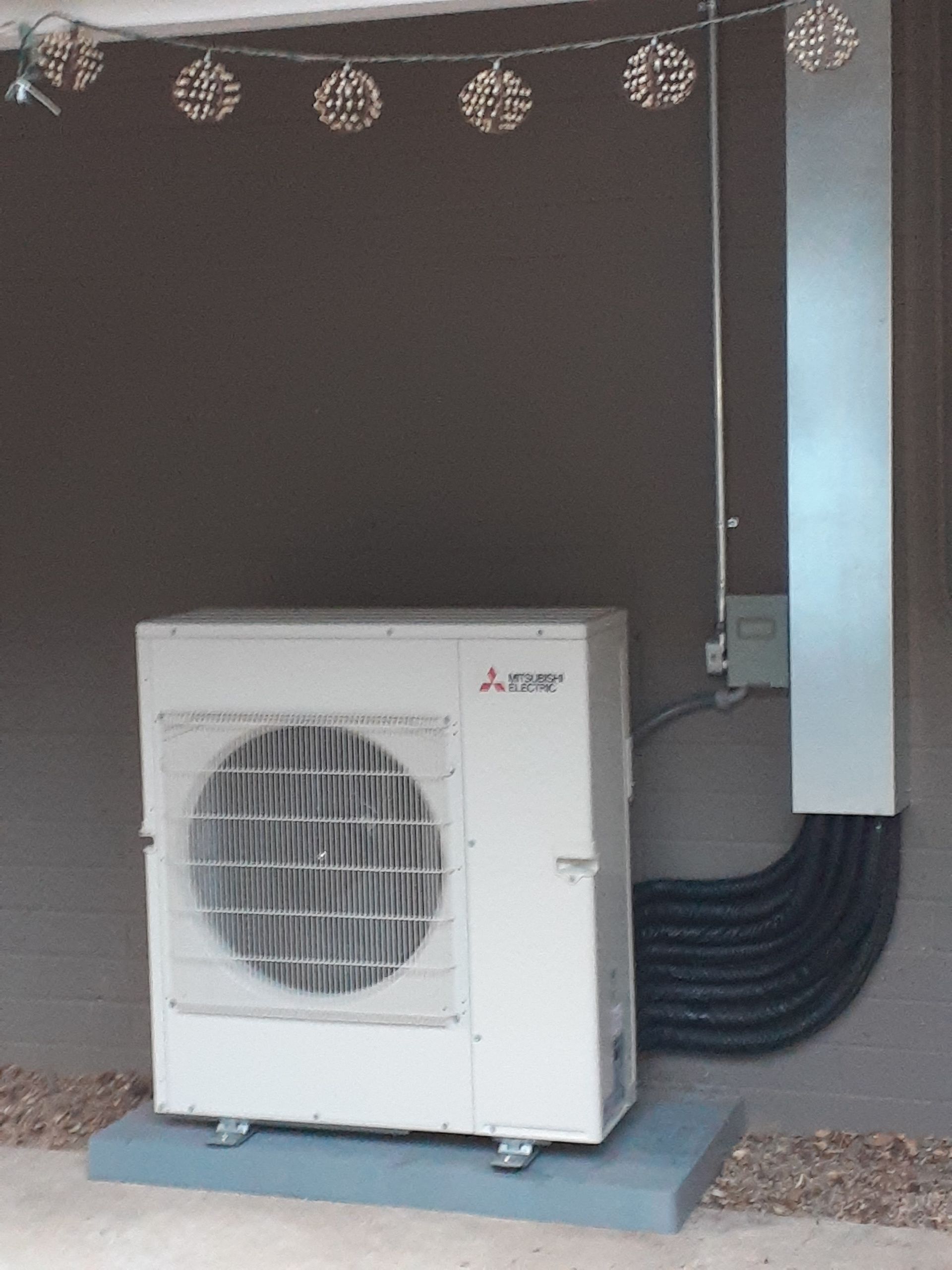 Outdoor air conditioning unit beside a building wall, with black hoses and decorative string lights above.