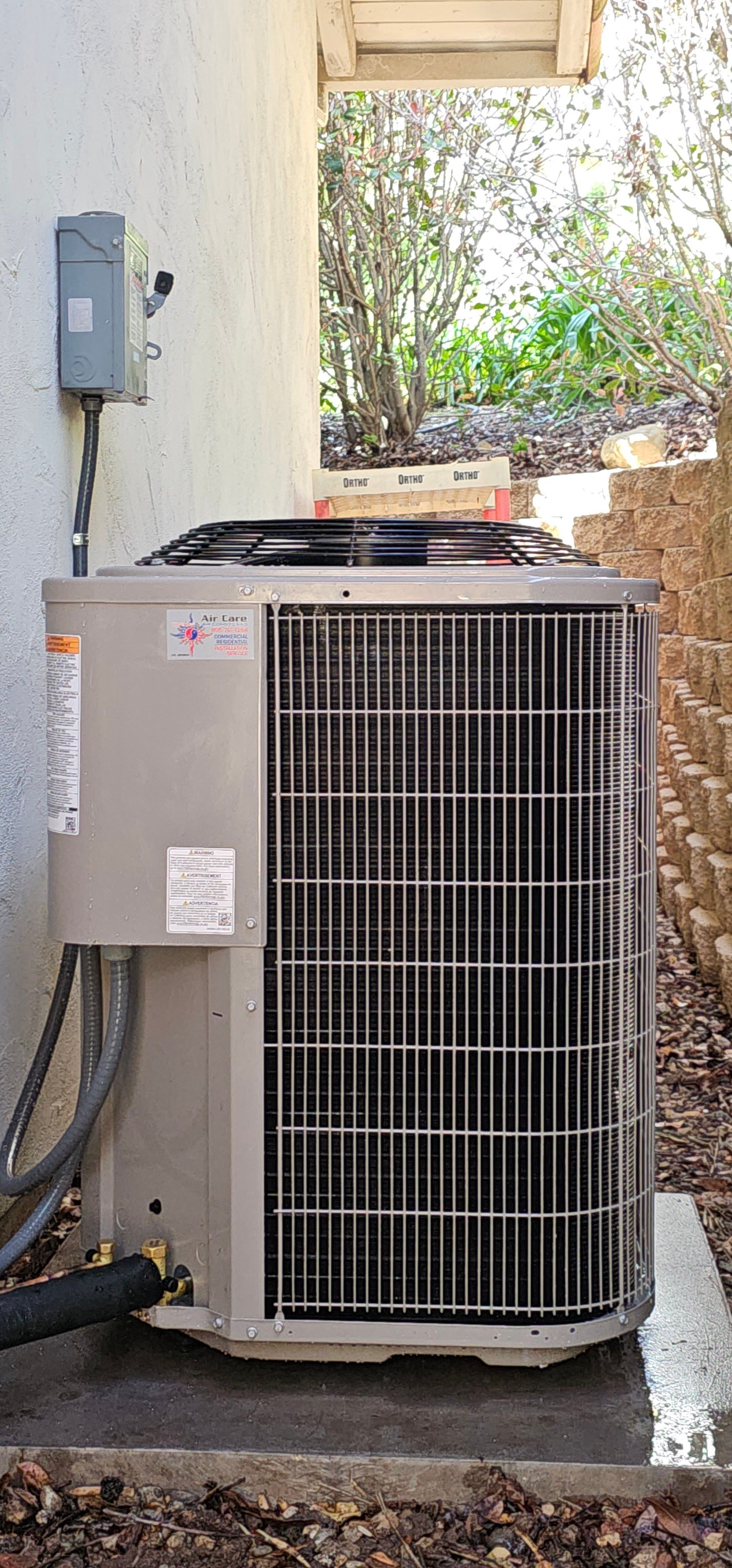 Outdoor HVAC condenser unit beside a house, with a utility meter on the wall and plants behind it