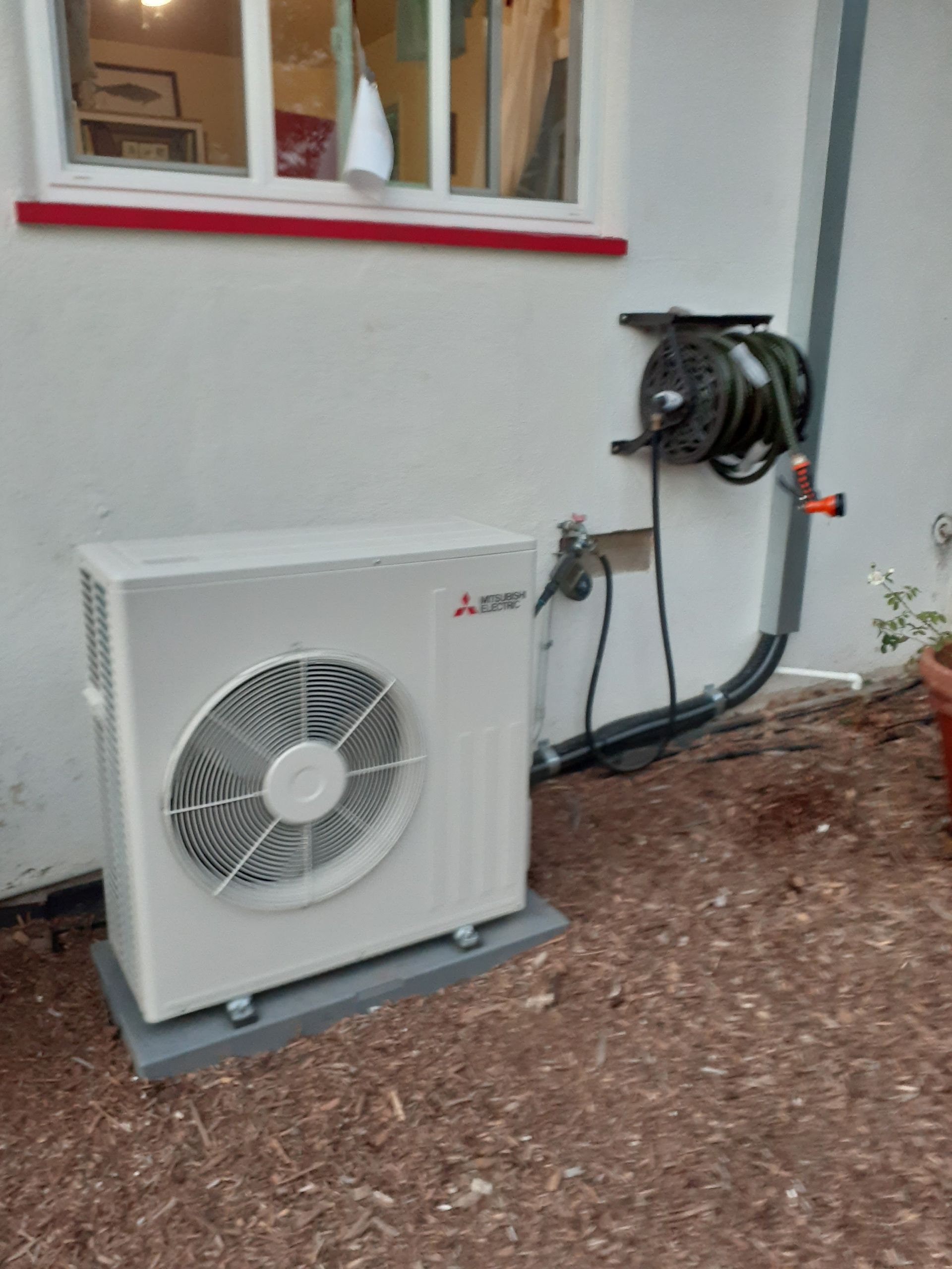 Outdoor HVAC unit beside a house wall, with a wall-mounted hose reel and window above.