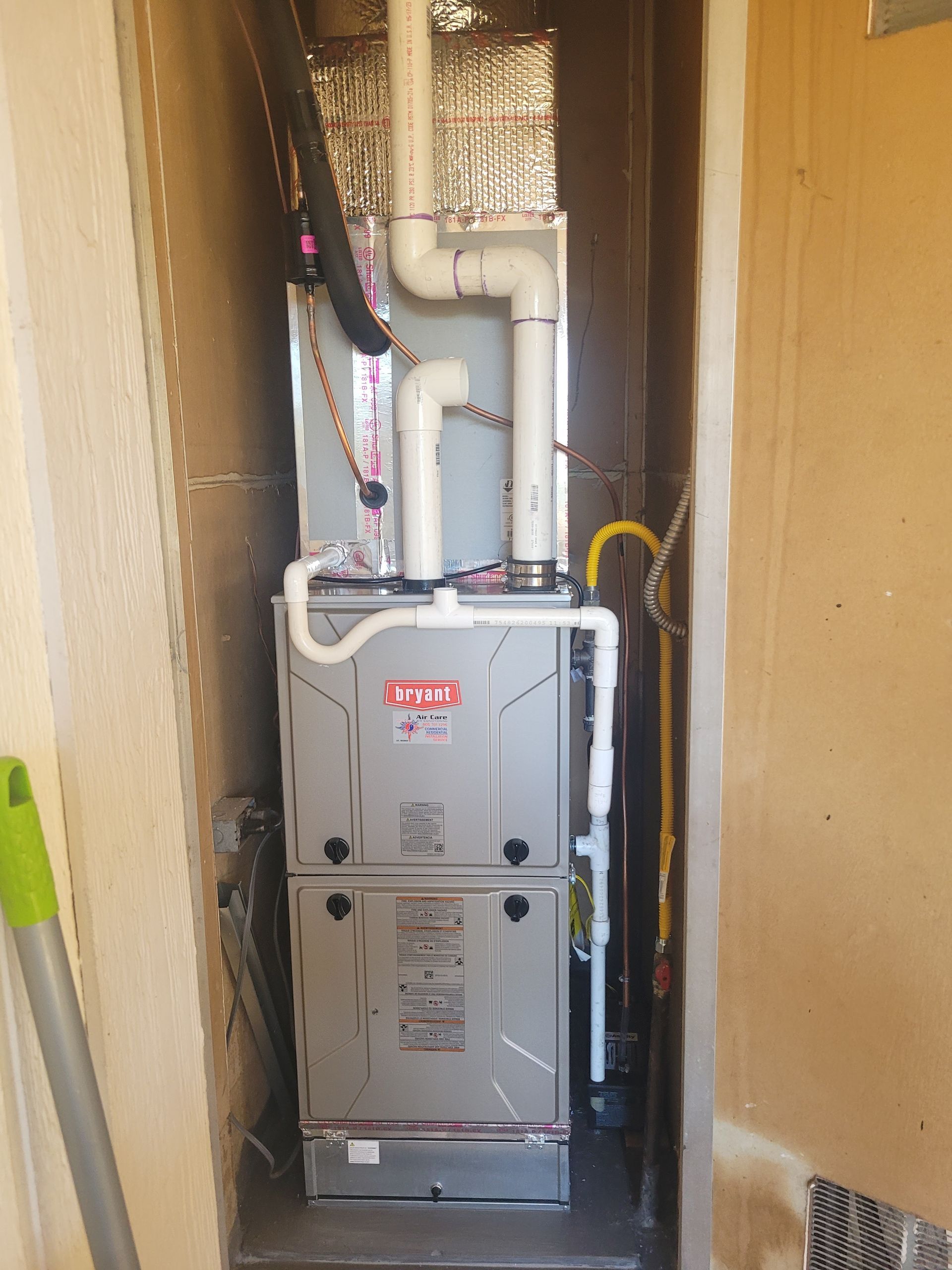 Gray furnace in a narrow closet with white ducting and pipes, viewed from the front.