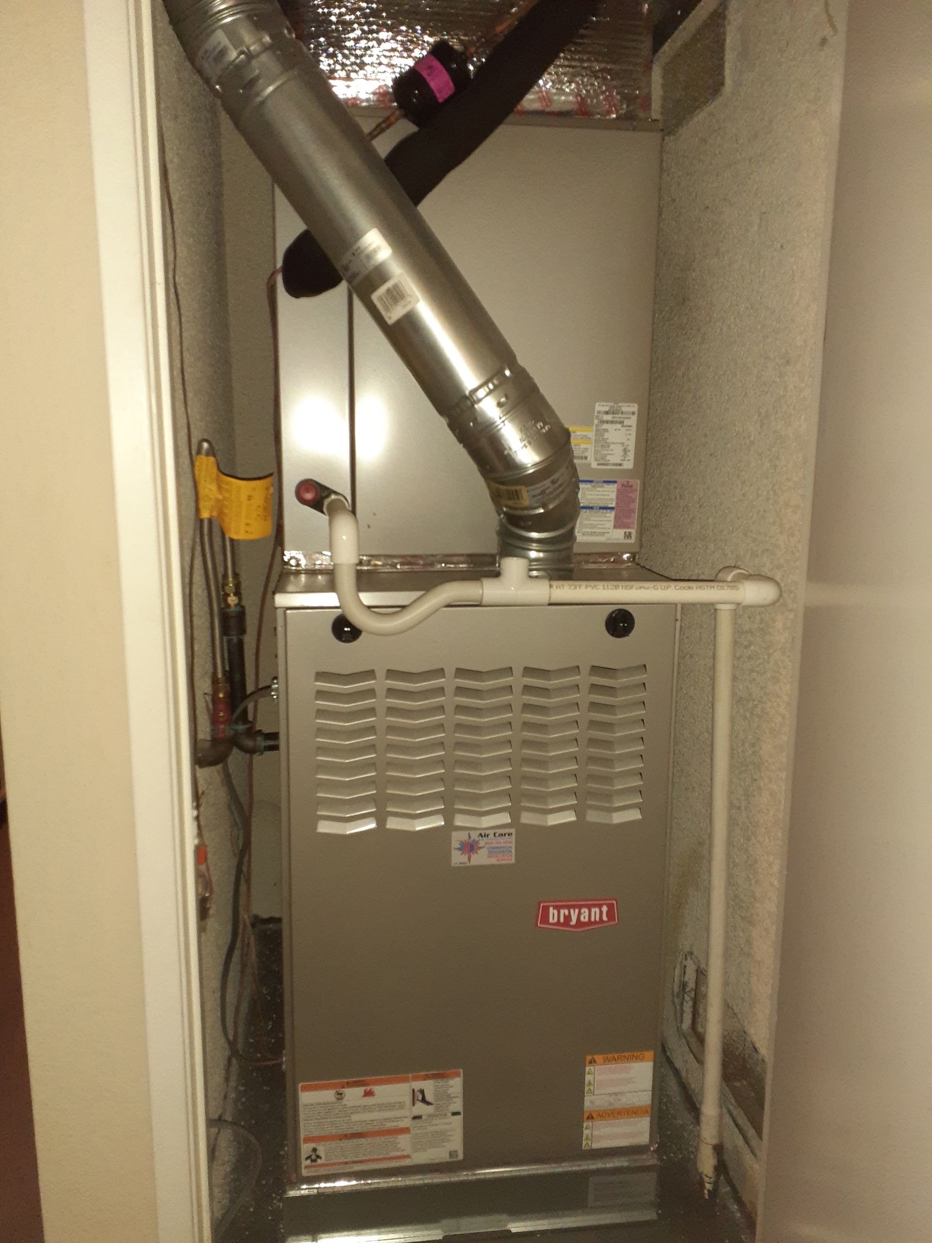 Furnace unit in a narrow utility closet with silver ductwork and pipes above it.