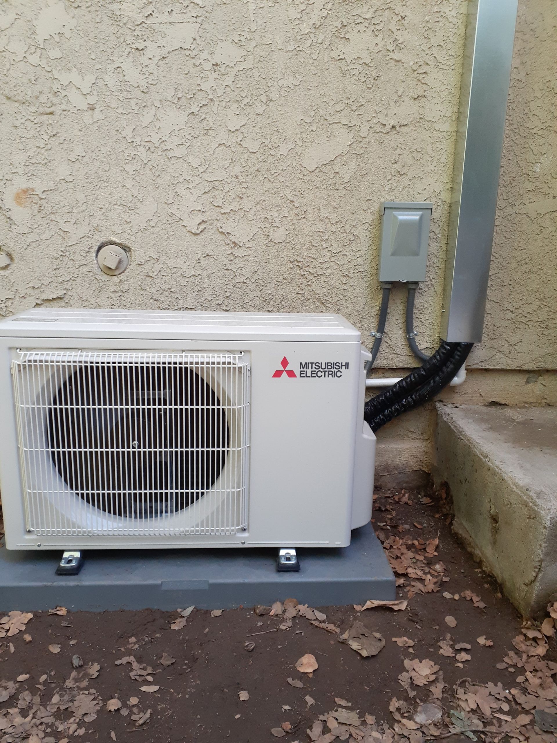 Outdoor Mitsubishi air conditioner unit beside a beige wall, with conduit and electrical box nearby.