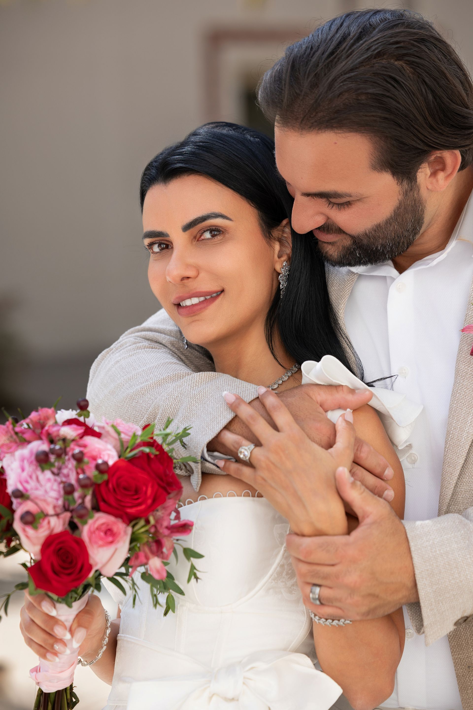 A man is hugging a woman in a wedding dress who is holding a bouquet of flowers.