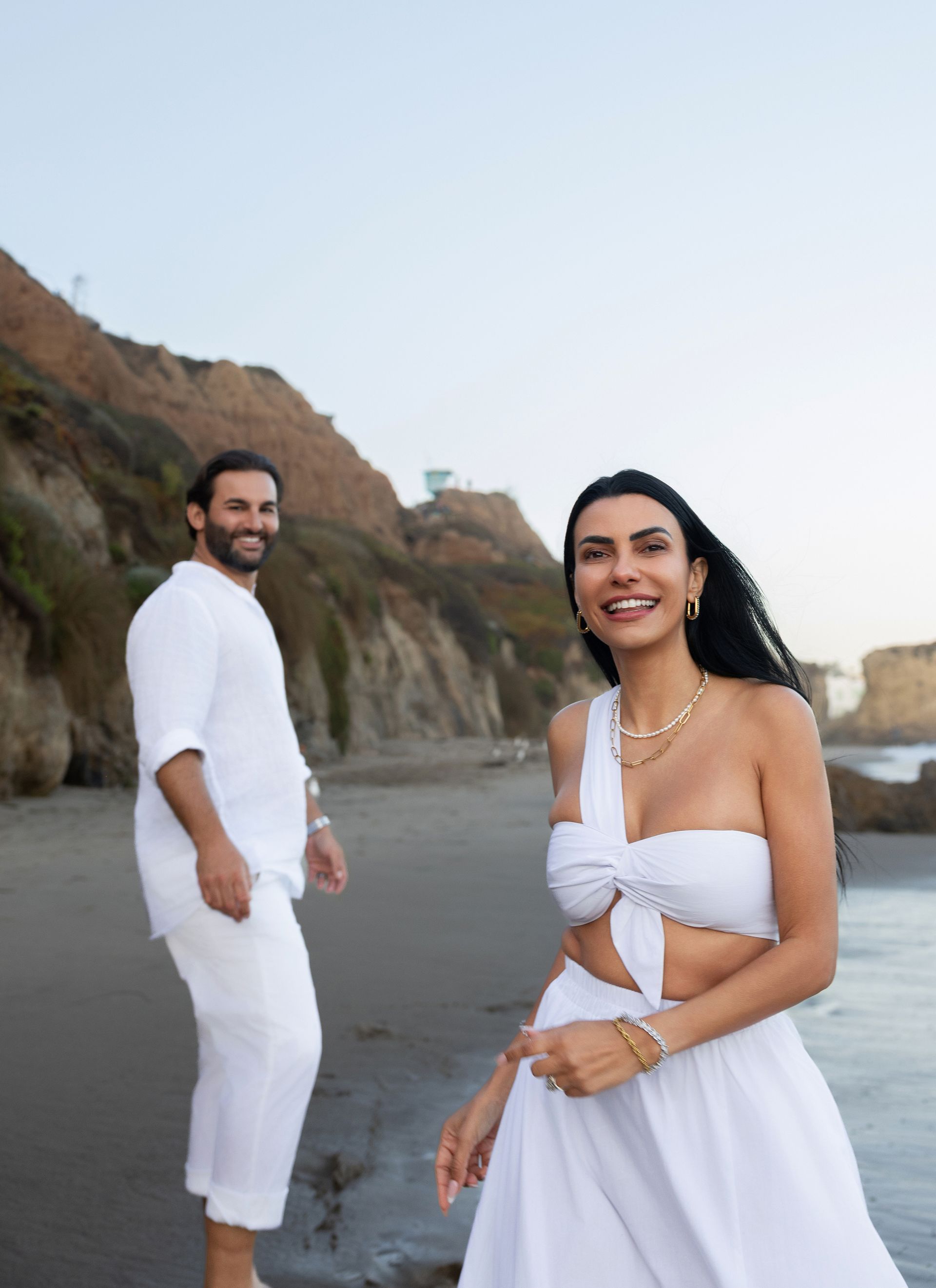 A man and a woman are standing on a beach . the woman is wearing a white dress.