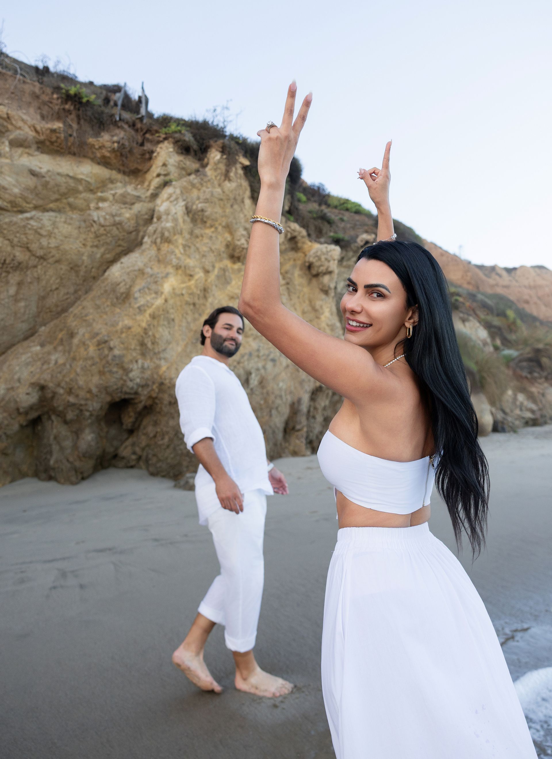 A man and a woman are standing on a beach . the woman is wearing a white dress.