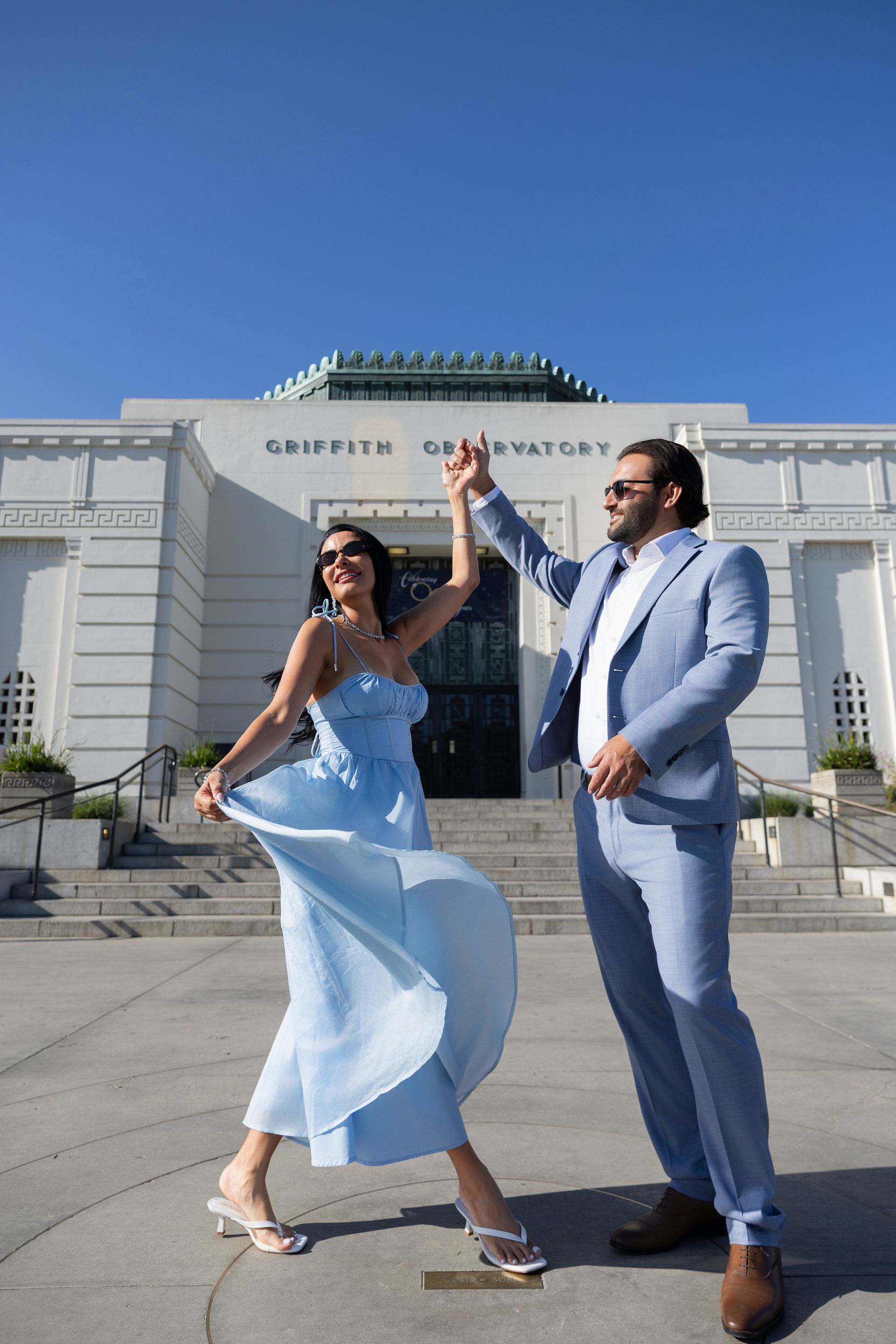 A man in a suit and a woman in a blue dress are dancing in front of a building.