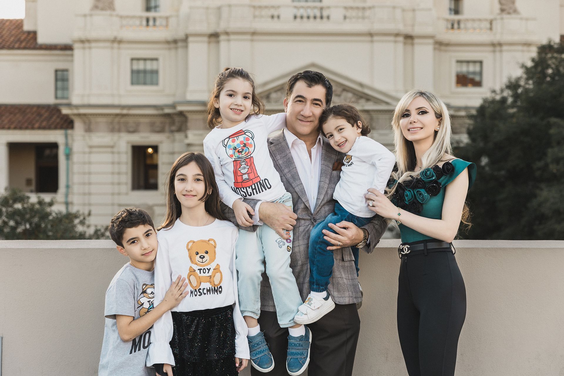 A family of five standing together for a portrait, with a warm and loving atmosphere. The family consists of a mother and father in their 30s, a teenage daughter, and two younger children (a son around 8 years old and a daughter around 5 years old). They are positioned in a semi-circle, with the parents in the middle, both smiling warmly. The teenage daughter stands to one side, slightly angled, while the two younger children are positioned in front of the parents, with the son sitting on the ground, and the younger daughter standing with her arms around her parents. The background features soft, natural lighting with a neutral-colored setting, perhaps a park or cozy indoor space. Everyone is dressed in casual but coordinated outfits, with light tones and complementary colors to create harmony in the image. The family members are all gazing at the camera with happy expressions