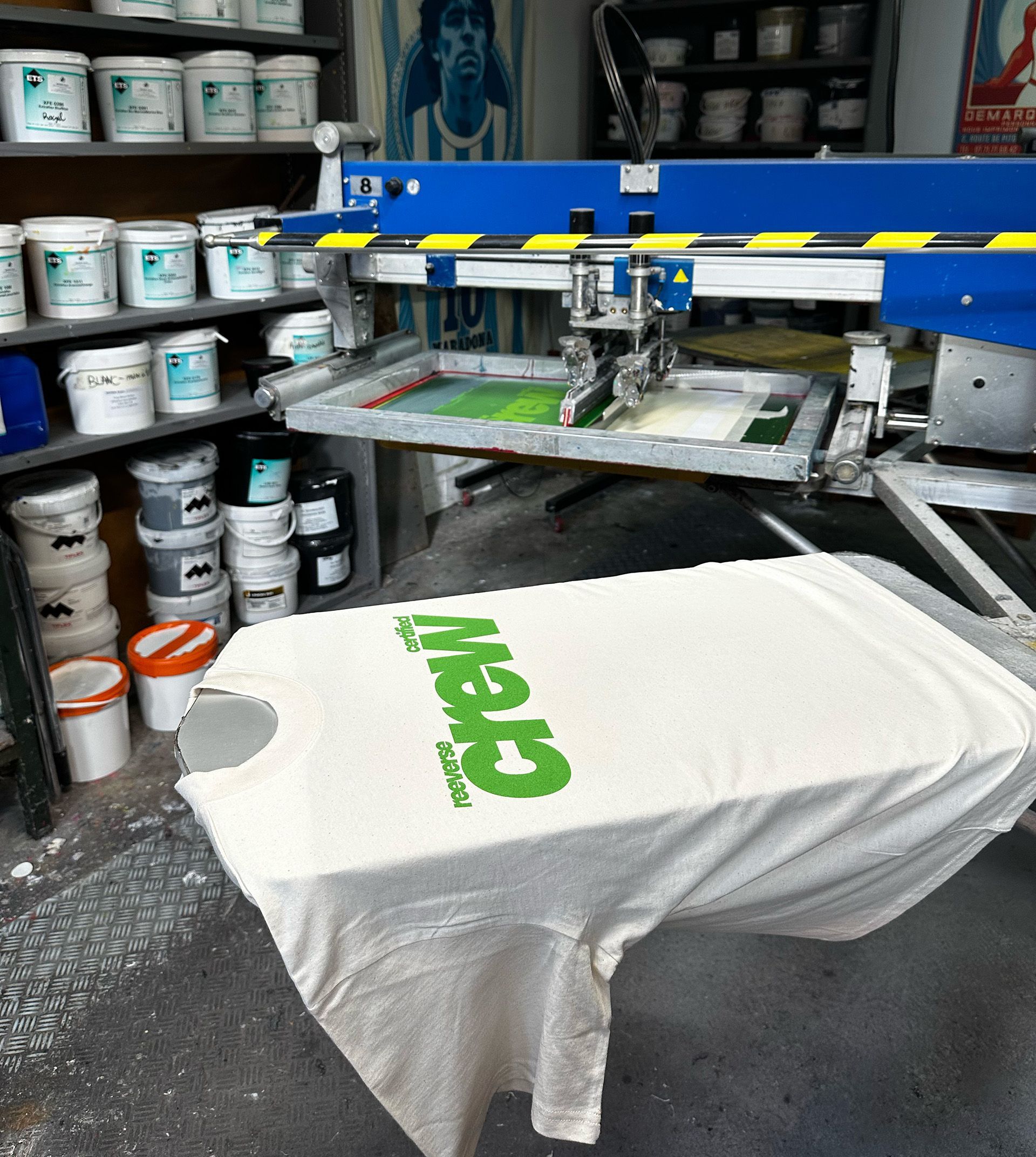 Un t-shirt blanc orné du logo vert « The Crew » est imprimé sur une machine de sérigraphie dans un atelier.