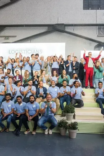 Un grand groupe de personnes en chemises bleu clair, assises sur des gradins, certaines debout et applaudissant, à l'intérieur d'un hall industriel.