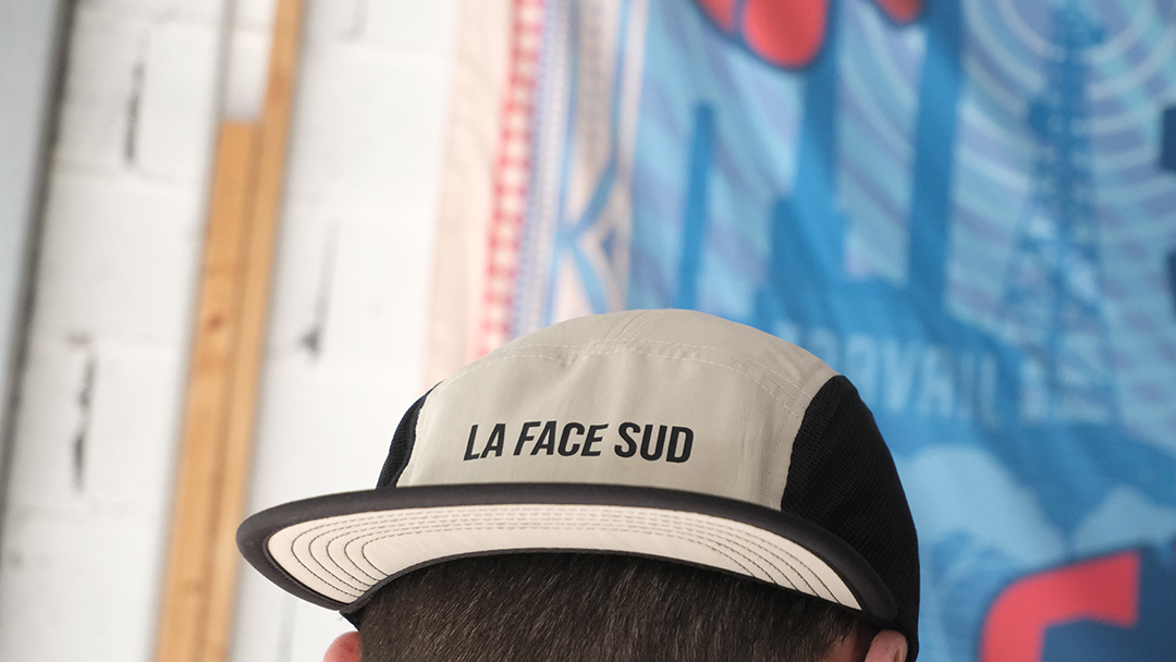 Une personne portant une casquette beige et noire à cinq panneaux avec l'inscription « LA FACE SUD » brodée sur le devant.
