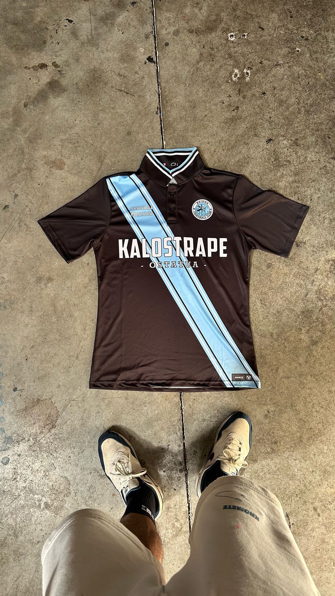 Un maillot de football marron avec une rayure diagonale bleu clair et des inscriptions blanches repose sur un sol en béton, au-dessus de vos pieds.