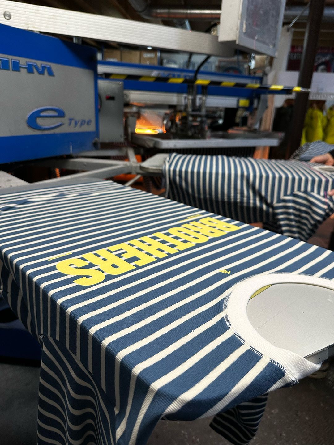 Une chemise à rayures bleues et blanches est posée sur une machine de sérigraphie, du texte jaune étant imprimé sur le tissu.