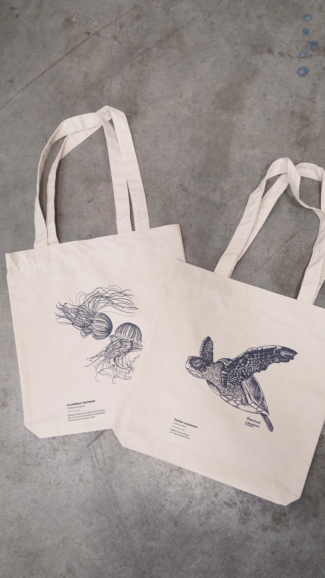 Deux sacs fourre-tout en toile beige clair, l'un orné d'une illustration de méduse et l'autre d'une tortue marine, posés sur du béton gris.