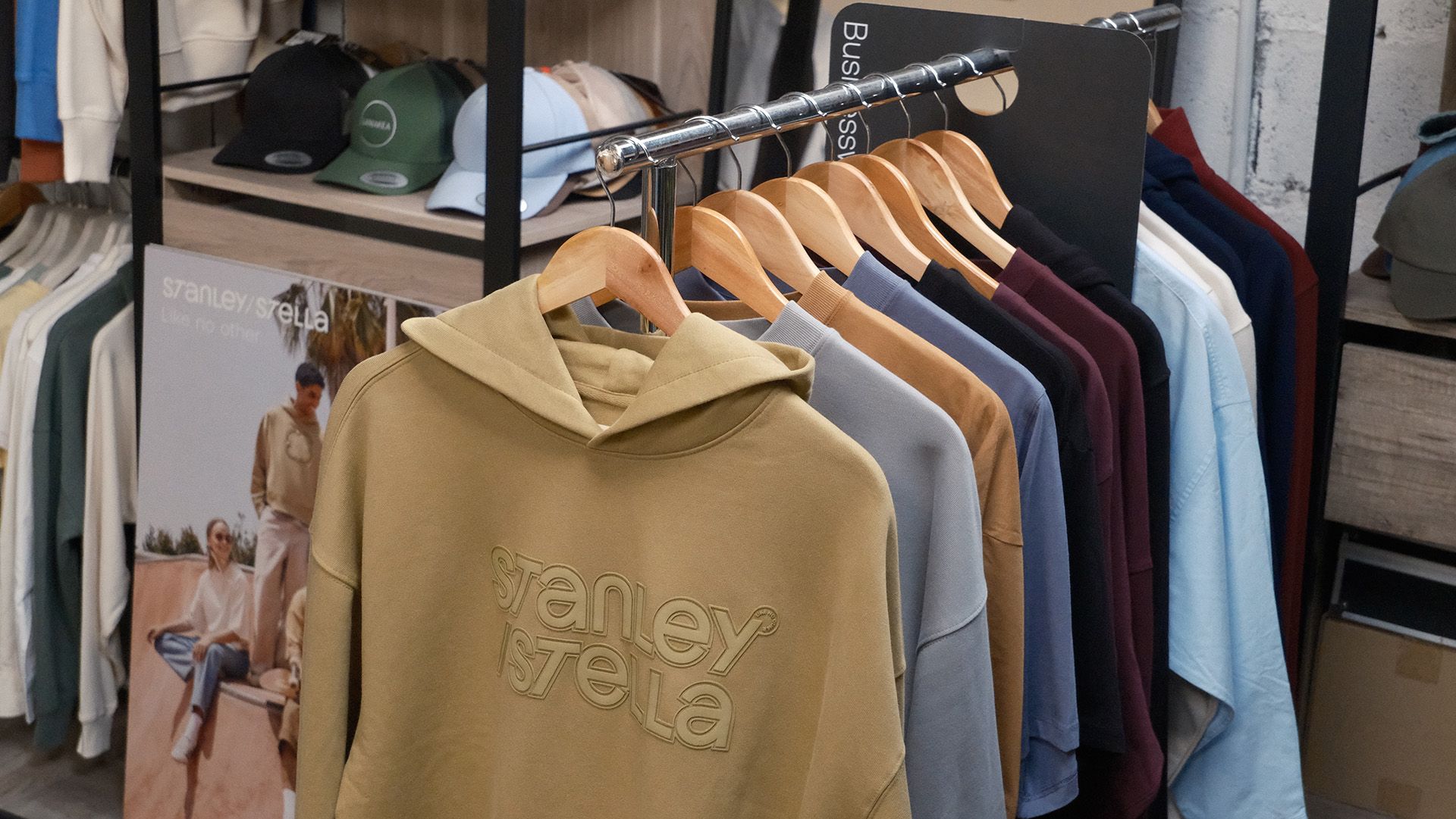 Un portant à vêtements accueille des sweats à capuche de différentes couleurs, notamment olive, bleu et noir, tandis que des casquettes pliées sont exposées sur une étagère au-dessus.