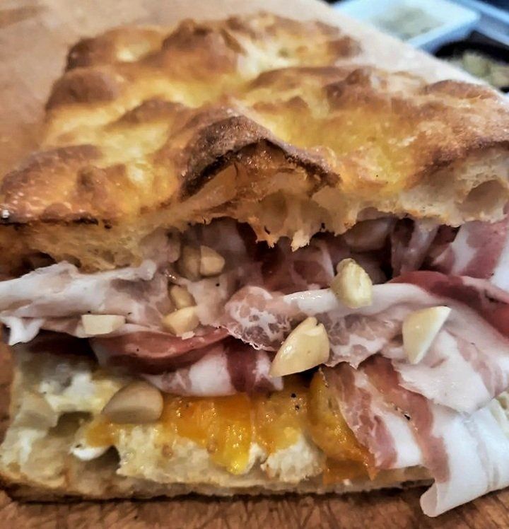 Un panino rustico su pane dorato e croccante, farcito con sottili fette di salumi, confettura d'arance e pinoli sparsi.