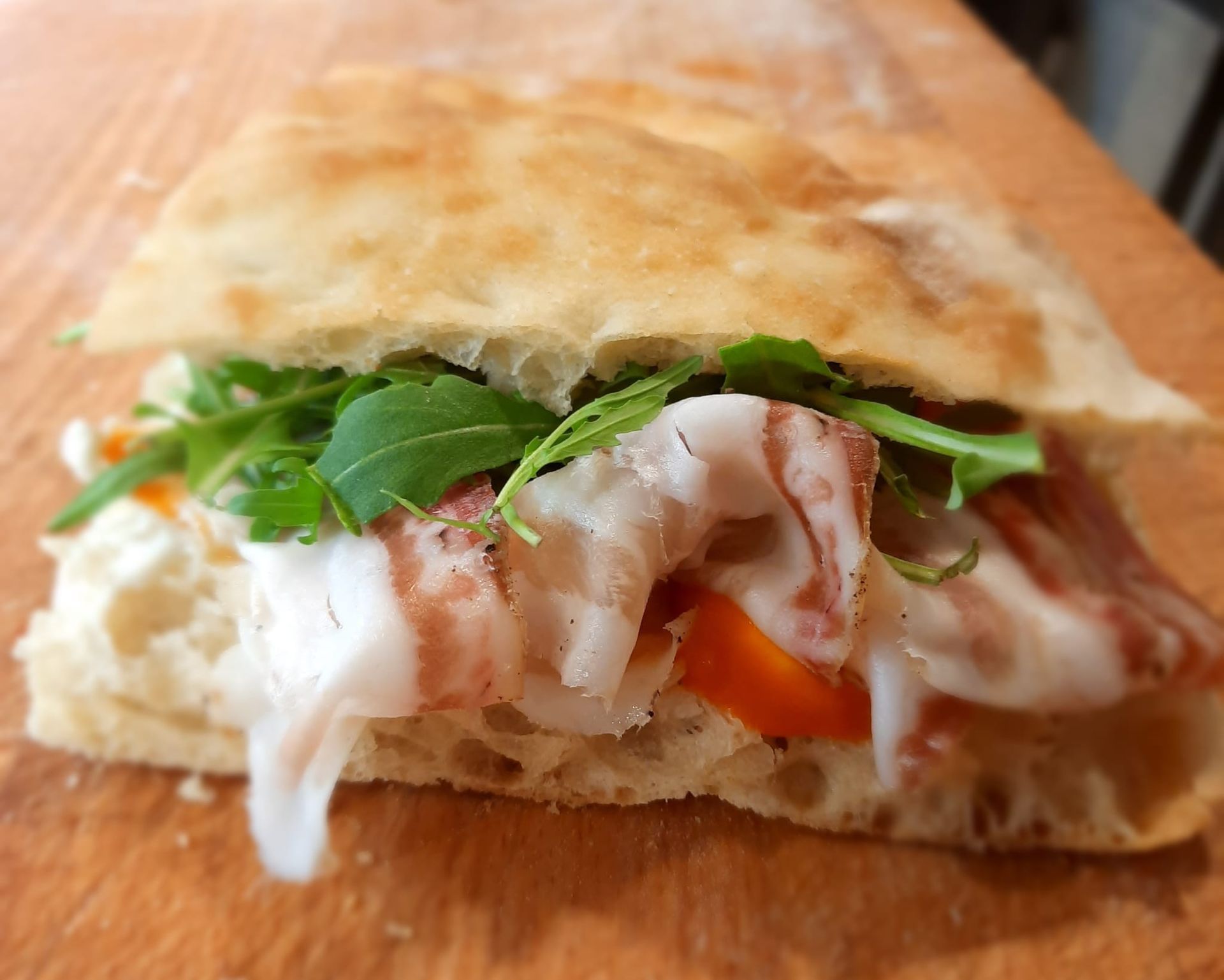 Un panino gourmet con salumi, rucola fresca e fette di pomodoro su una focaccia tostata a mollica aperta, servita su una superficie di legno.