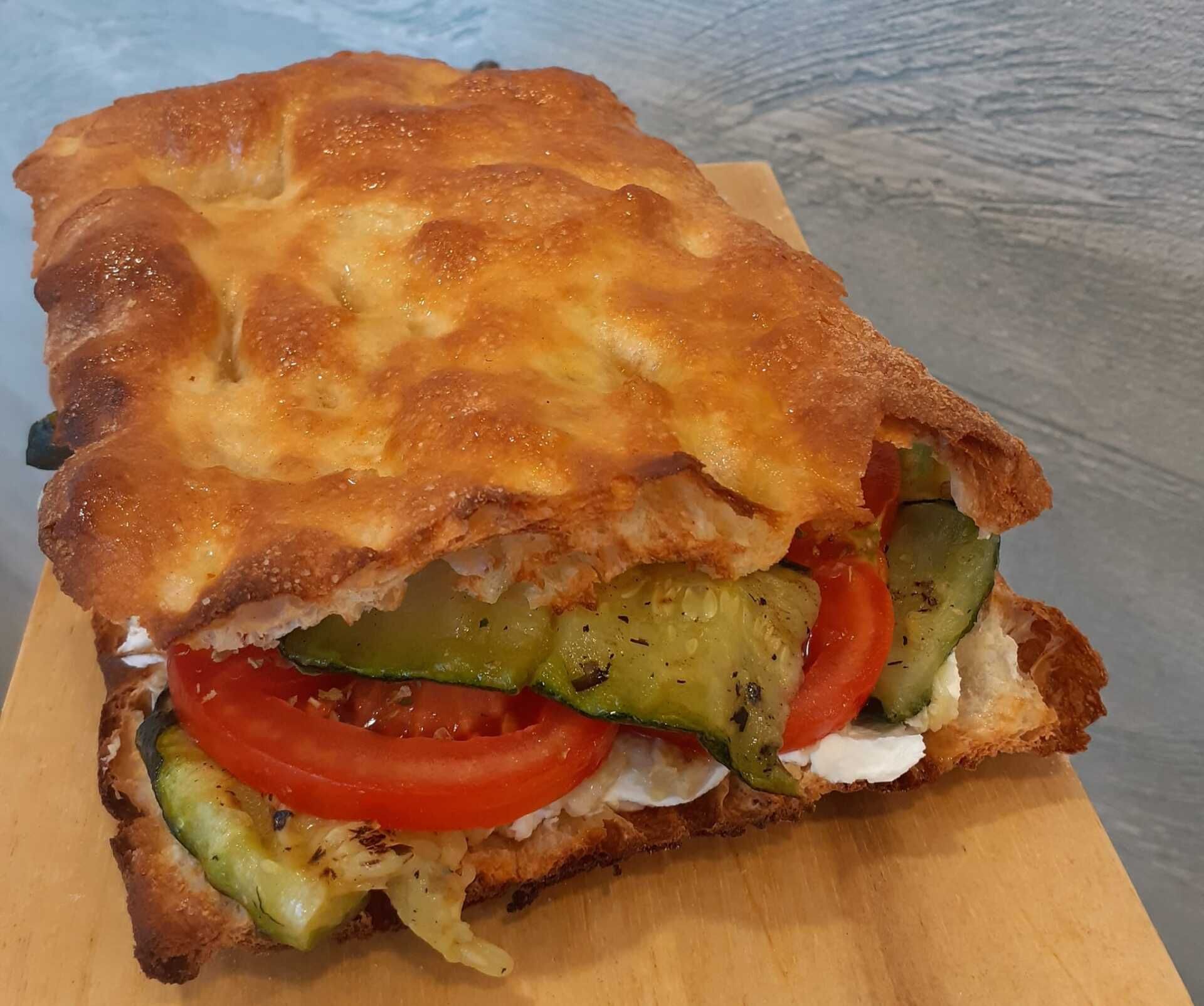 Un panino su focaccia dorata farcito con pomodoro fresco, zucchine grigliate e formaggio bianco cremoso, servito su un tagliere di legno.