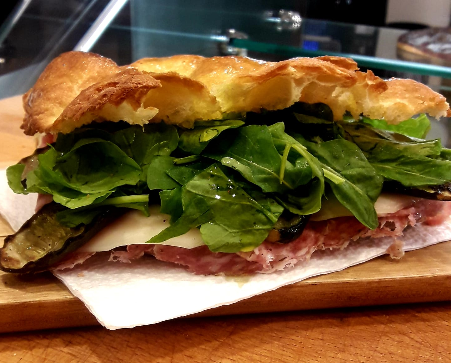 Un panino rustico su un tagliere di legno, farcito con verdure a foglia verde, affettati, formaggio e melanzane grigliate.