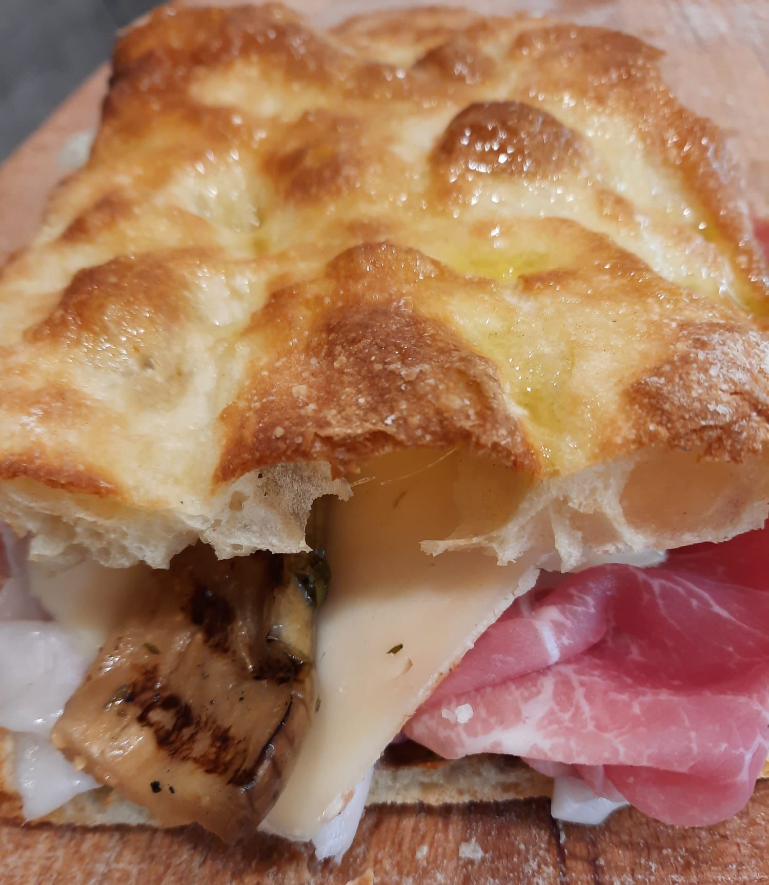 Un panino di focaccia con prosciutto crudo a fette, una fetta di formaggio e pezzi di funghi grigliati.