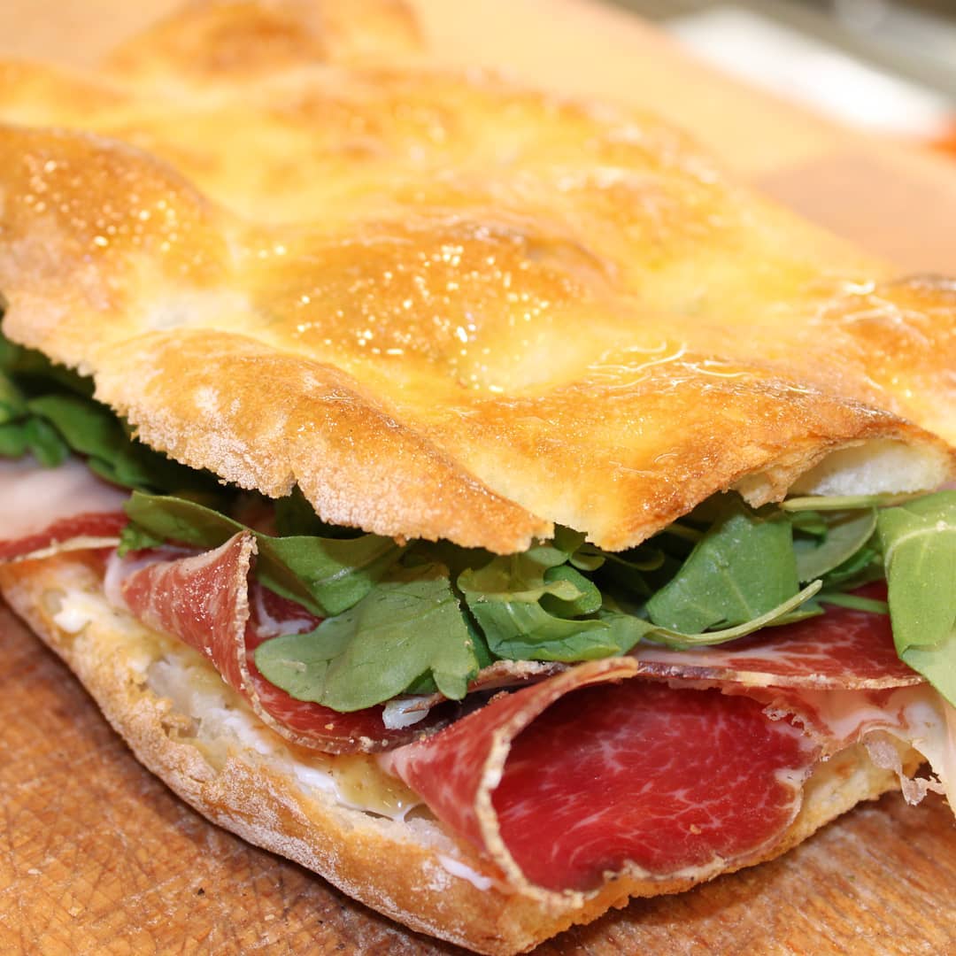 Primo piano di un panino con affettati sottili e rucola fresca su una focaccia croccante e dorata.