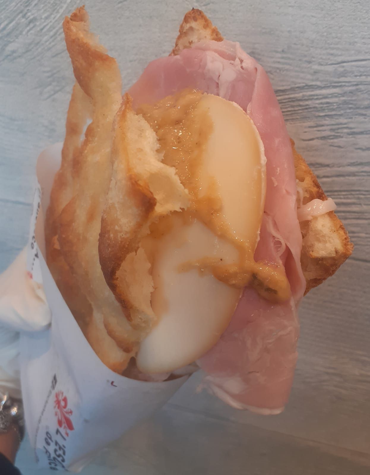 Un panino avvolto in carta, con prosciutto cotto piegato e fette di formaggio bianco con una salsa dorata su pane tostato.