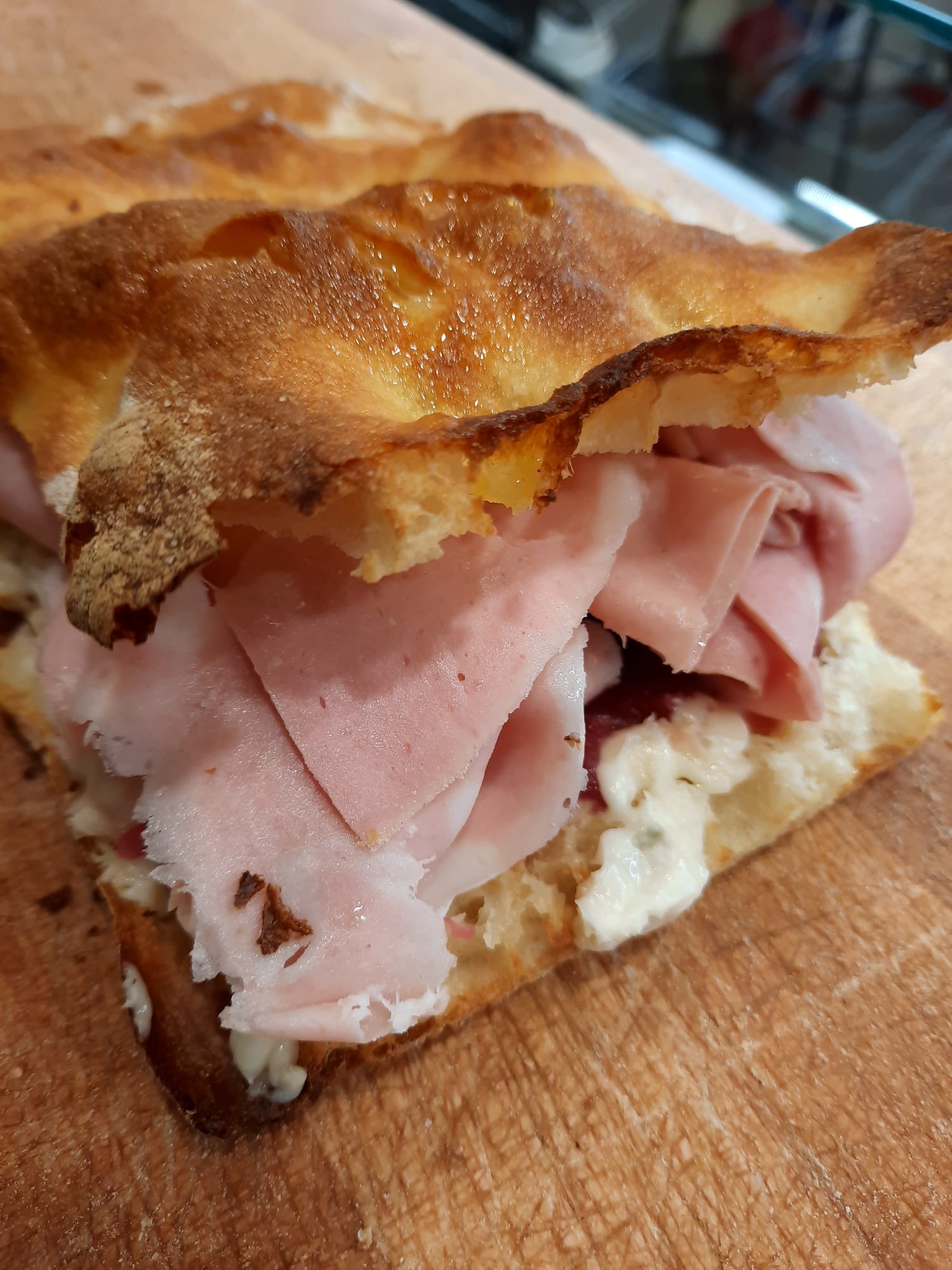 Un panino con focaccia croccante e dorata, farcito con prosciutto a fette sottili e formaggio bianco cremoso, servito su una superficie di legno.