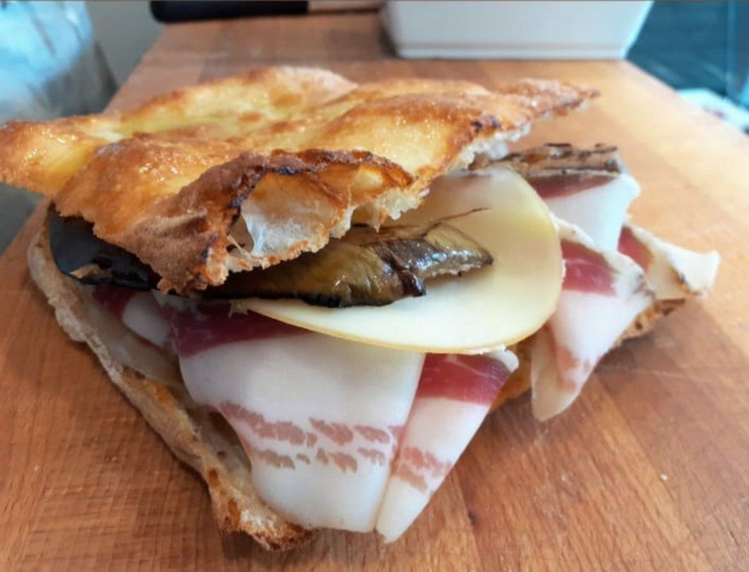 Un panino su un tagliere di legno farcito con sottili fette di salumi, una fetta di formaggio e melanzane grigliate.