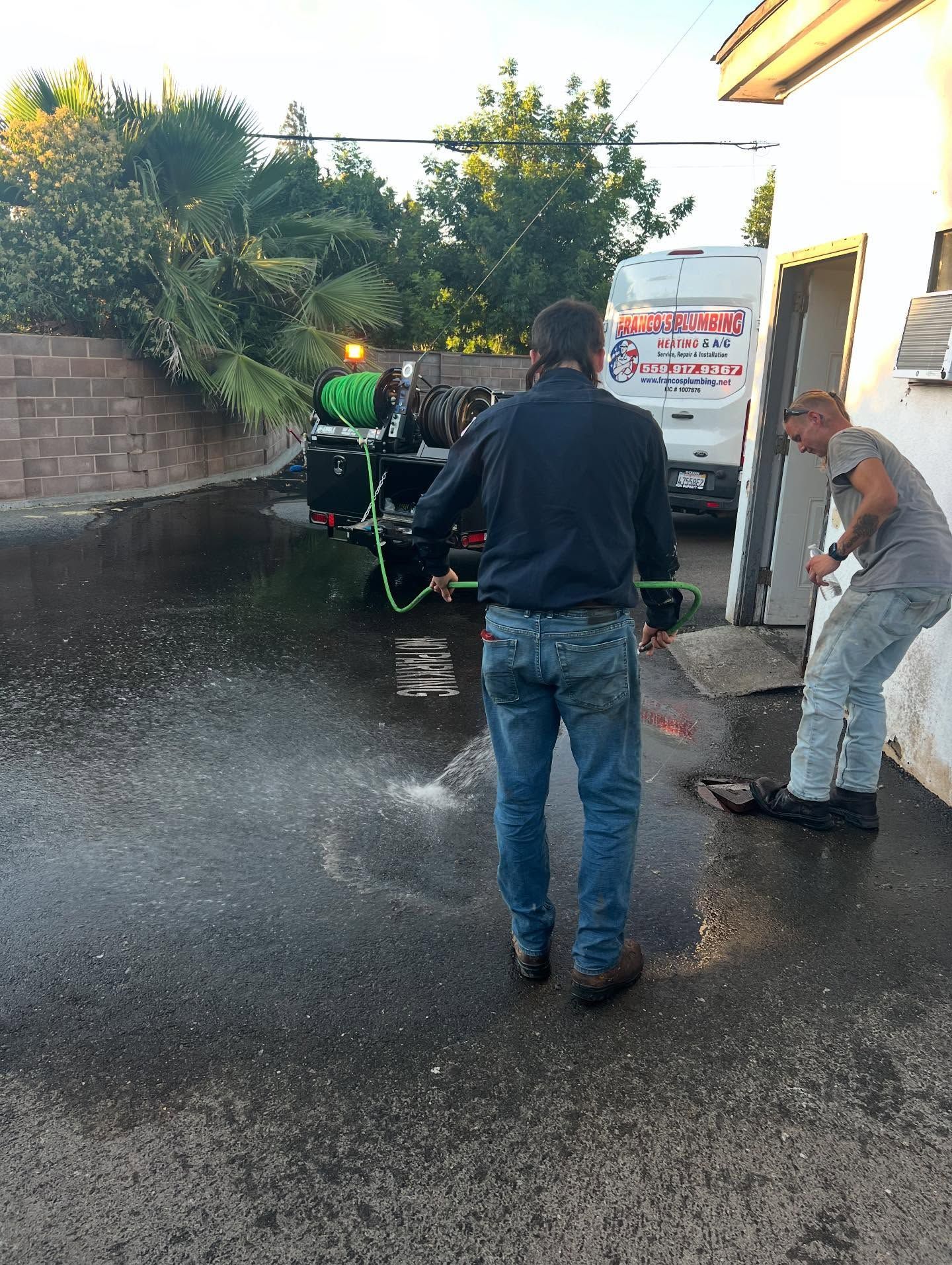 Two men pressure washing a wet paved area. One man holds the hose, while the other works on a building wall.