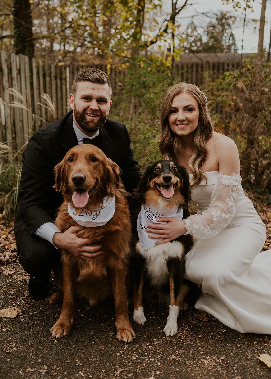 Couple in formalwear seated outdoors with two dogs in a wooded setting
