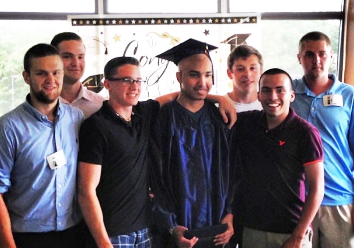 Six smiling people posing indoors, one wearing a graduation cap and gown.