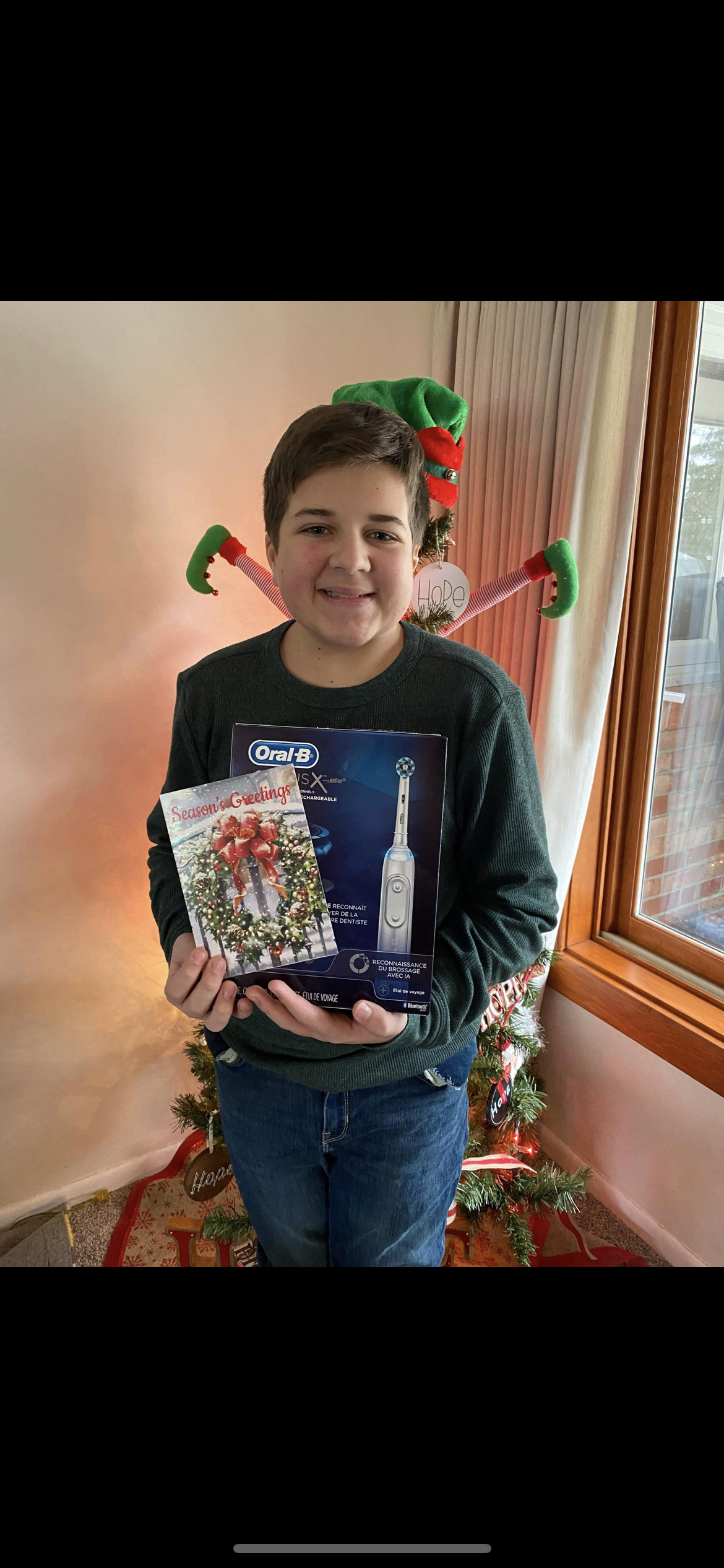 Person smiling indoors, holding a wrapped gift box with a festive tree in the background