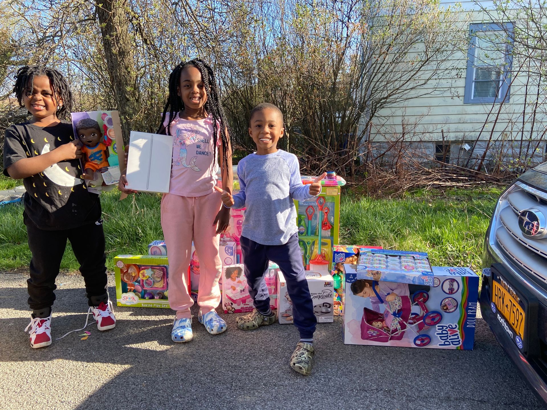 Three children standing outdoors with toys and gift boxes beside a car and house