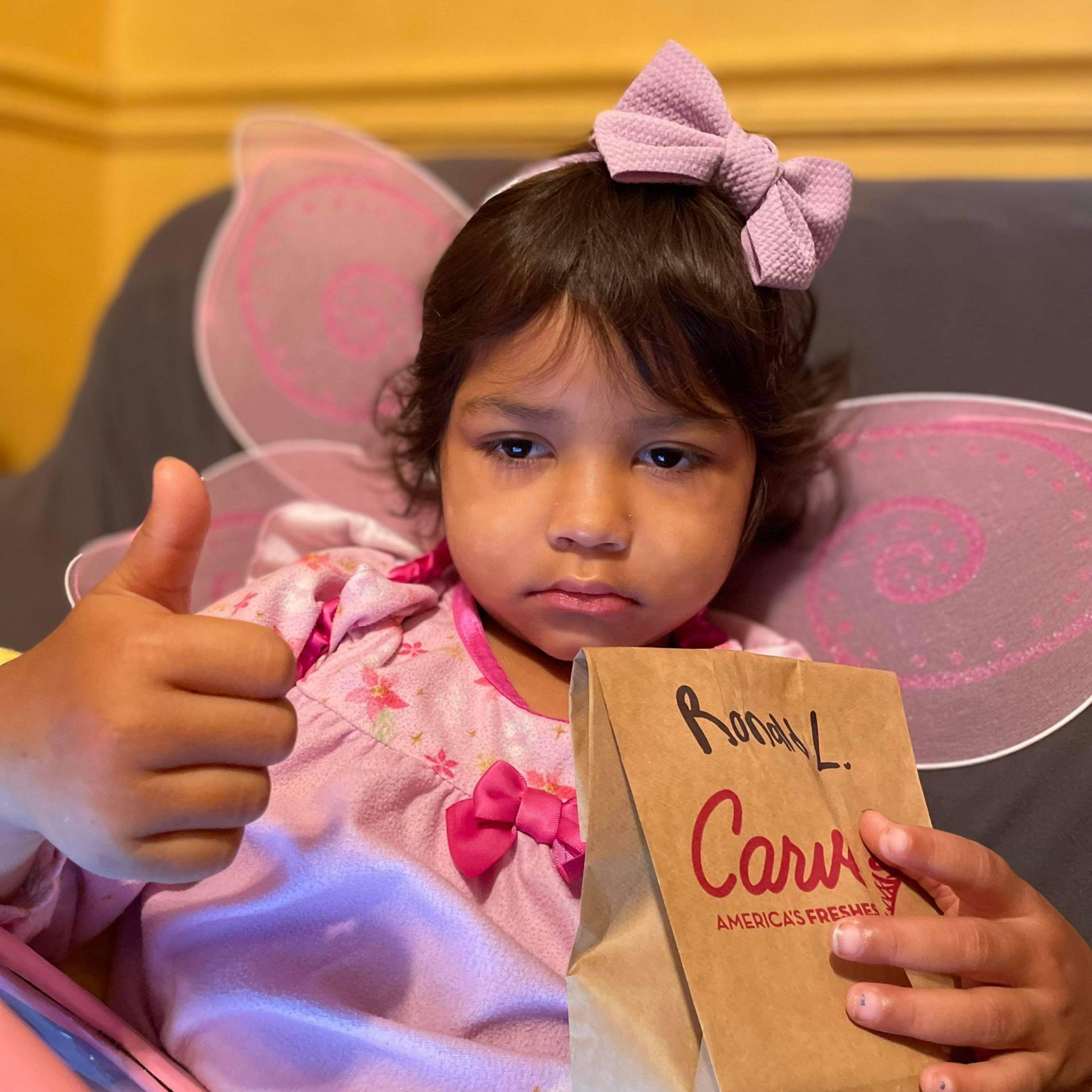 Child in pink outfit with butterfly wings gives a thumbs-up while holding a wrapped gift box.