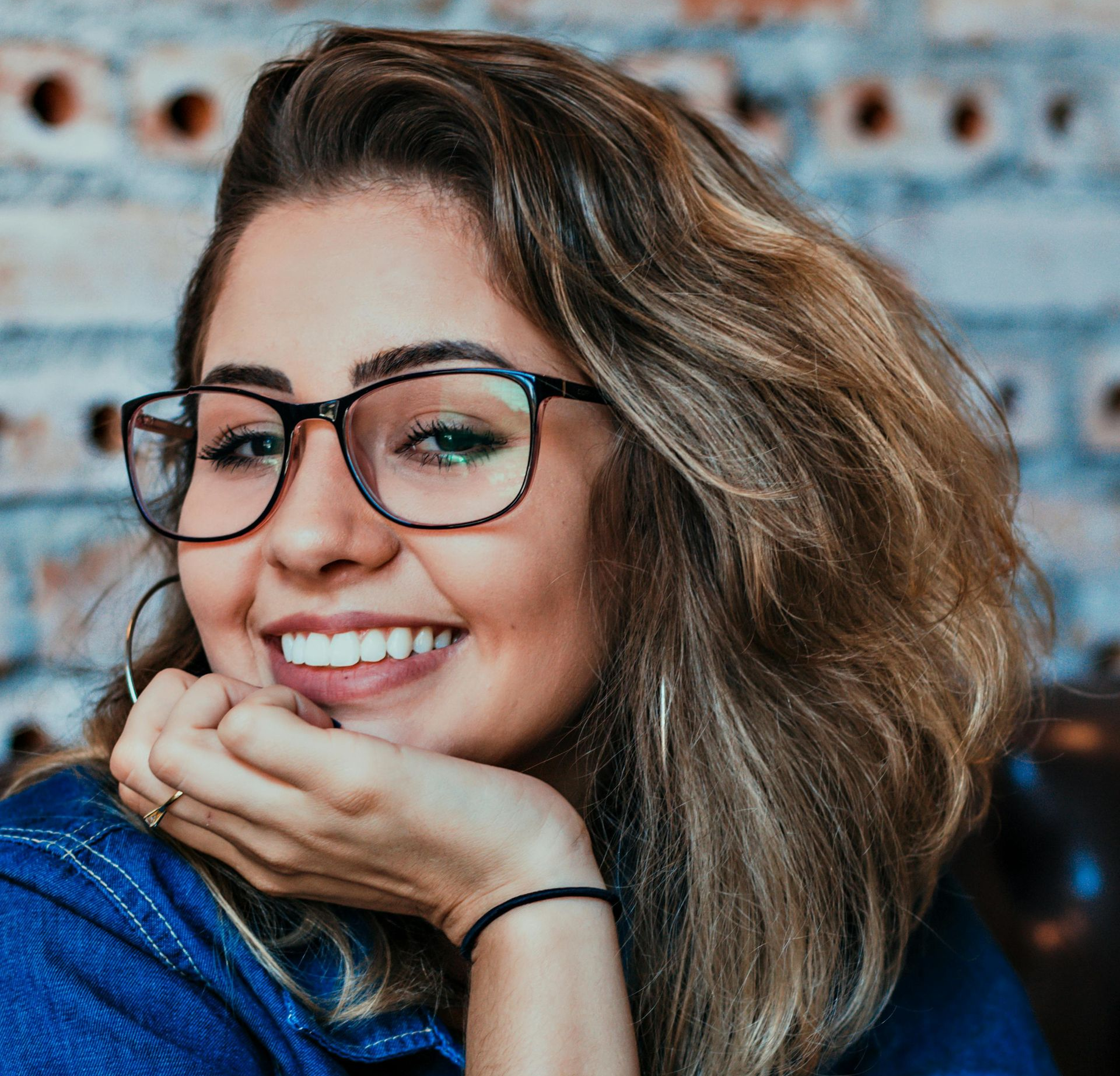 Smiling person with glasses resting chin on hand against a blurred blue background