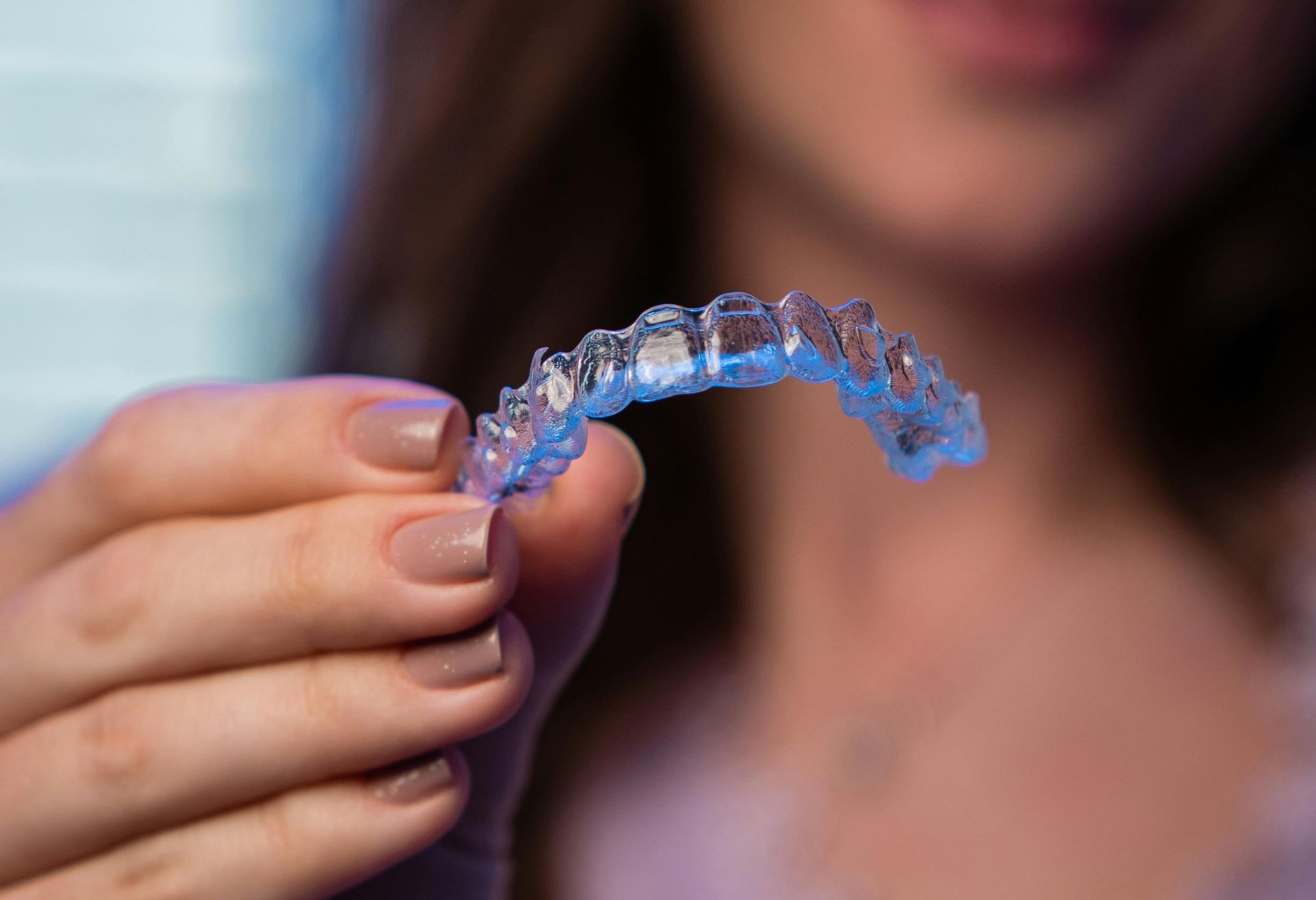A hand holding a clear dental aligner close to the camera, with a blurred woman in the background.