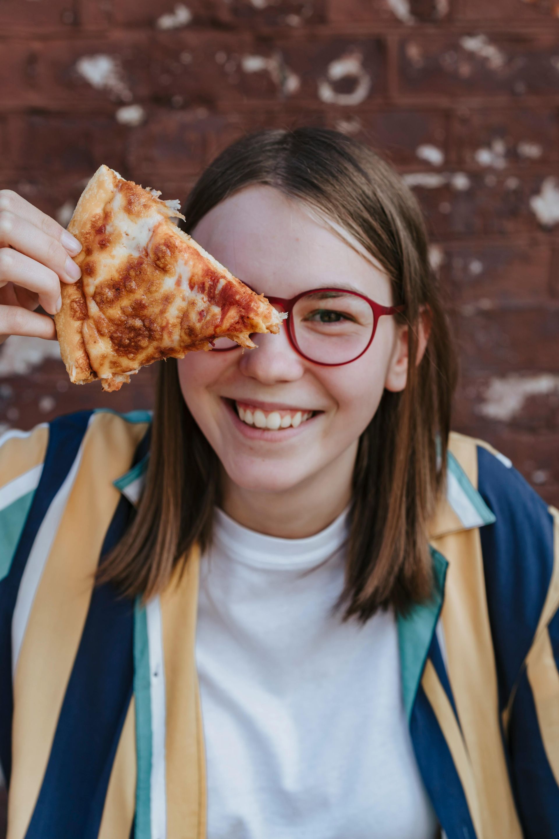 Smiling person holding a slice of pizza in front of one eye, wearing red glasses against a brick wall