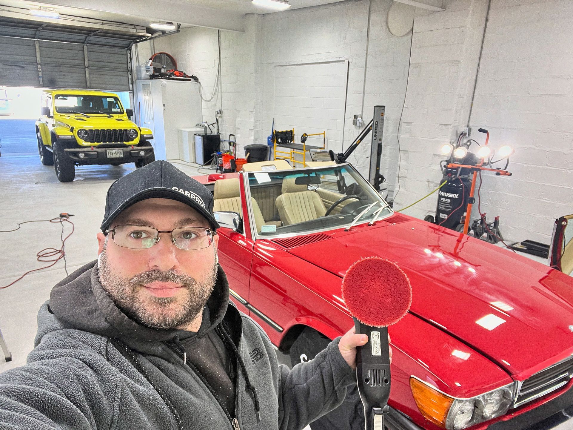 Man in a garage taking a selfie beside a red convertible, with a yellow truck in the background
