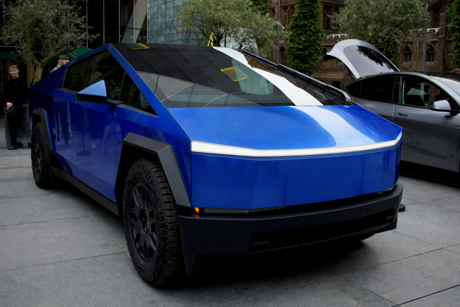 Blue Tesla Cybertruck parked on a street beside a gray car and greenery