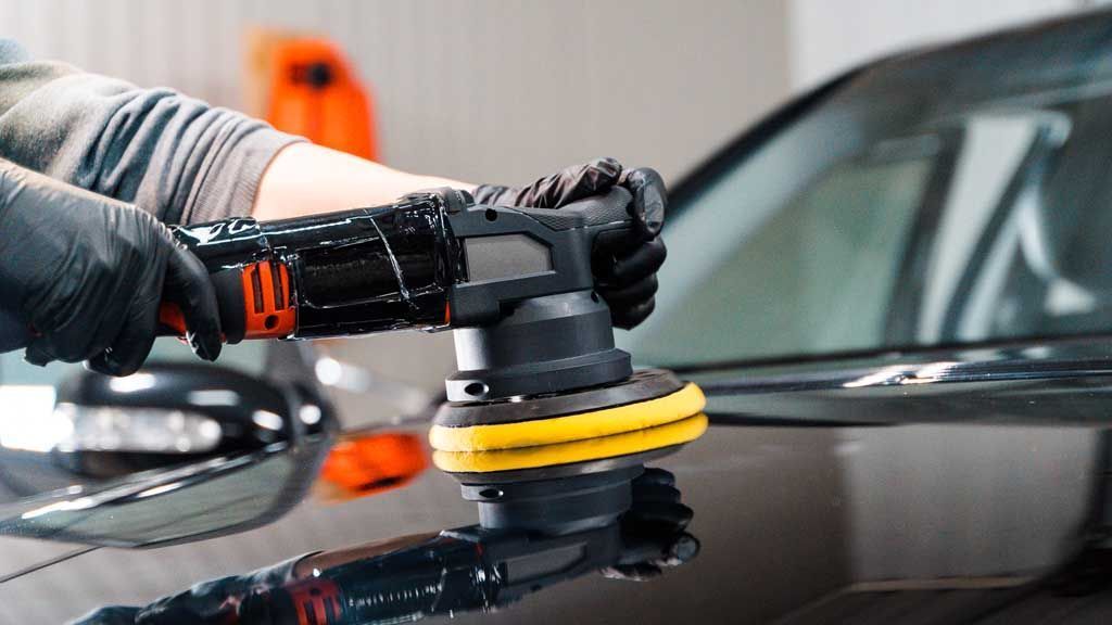 Gloved hands polishing a black car with a power buffer in a garage