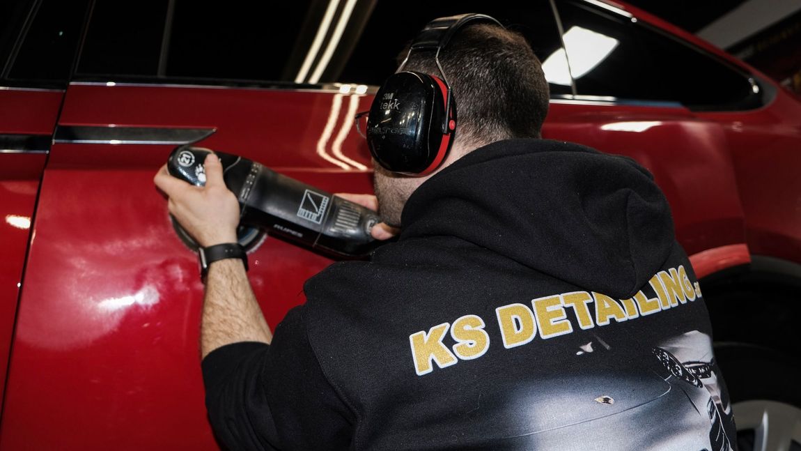 Detailer polishing a red car with a black machine polisher, wearing headphones and a KS Detailing jacket