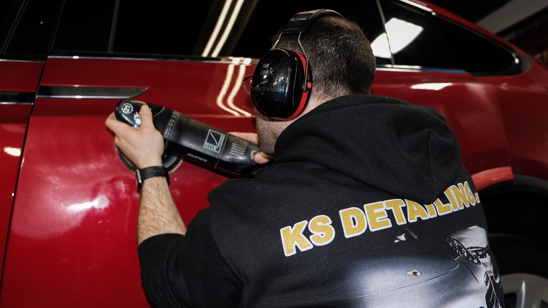 Detailer polishing a red car with a black machine polisher, wearing headphones and a KS Detailing jacket